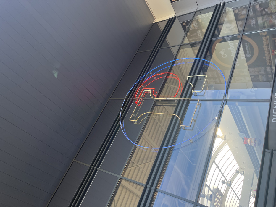 The image shows a large, modern building with a glass facade in Saint-Brice-Courcelles, France, a suburb of Reims. The building has a large, neon sign in the form of a circle with a letter "T" inside it. The neon is red, yellow, and blue. Behind the sign, a glass roof with a white steel structure and an array of large light fixtures can be seen. There are other large, modern signs with unintelligible text in the background.