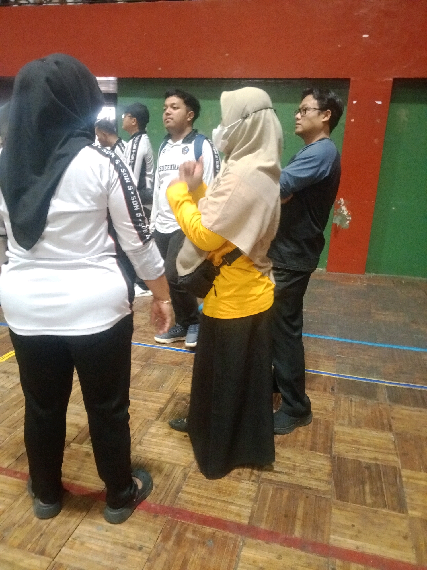 The image captures an indoor scene, likely a sports hall, with a wooden parquet floor marked with blue and red lines. The background features a wall painted in sections of orange and green. Several individuals are present, dressed casually. In the foreground, a person wearing a black hijab and white long-sleeved shirt with black pants is facing away from the camera. To their right, another person in a light brown hijab and yellow long-sleeved shirt with black pants is also visible, wearing a black bag across their chest. Further back, several other individuals are seen, including a man wearing glasses and a blue shirt. The environment suggests a gathering or event taking place in a public facility. Without further context, the specific location in Wuryorejo, Indonesia, cannot be determined from the visual information alone.