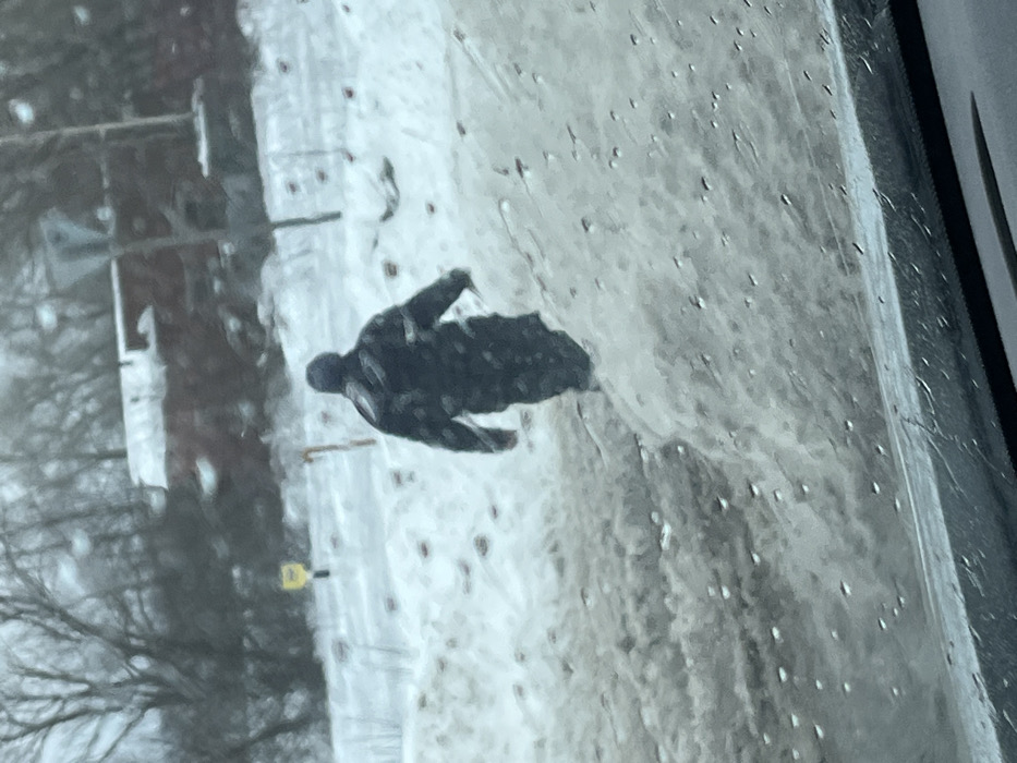 The image depicts a winter scene in Rogachovo, Russia, observed from inside a vehicle through a wet windshield. A single individual, viewed from behind, is walking away from the camera through snow-covered terrain. The person is dressed in dark, possibly patterned, winter outerwear including a hooded jacket or coat and a dark beanie, and appears to be holding a long, thin object in their right hand, pointed towards the ground. The immediate foreground shows a significant accumulation of dirty, partially melted snow and slush on the right side, along with the lower edge of the vehicle's interior. The ground where the person is walking is covered in snow, with visible darker patches possibly indicating exposed earth or debris. In the mid-background, a small yellow sign is visible to the left, and a utility pole or similar structure stands to the right. The far background features leafless trees and a reddish-brown building with a snow-covered roof, partially obscured by the trees. The overall environment is overcast and appears cold, consistent with winter conditions.