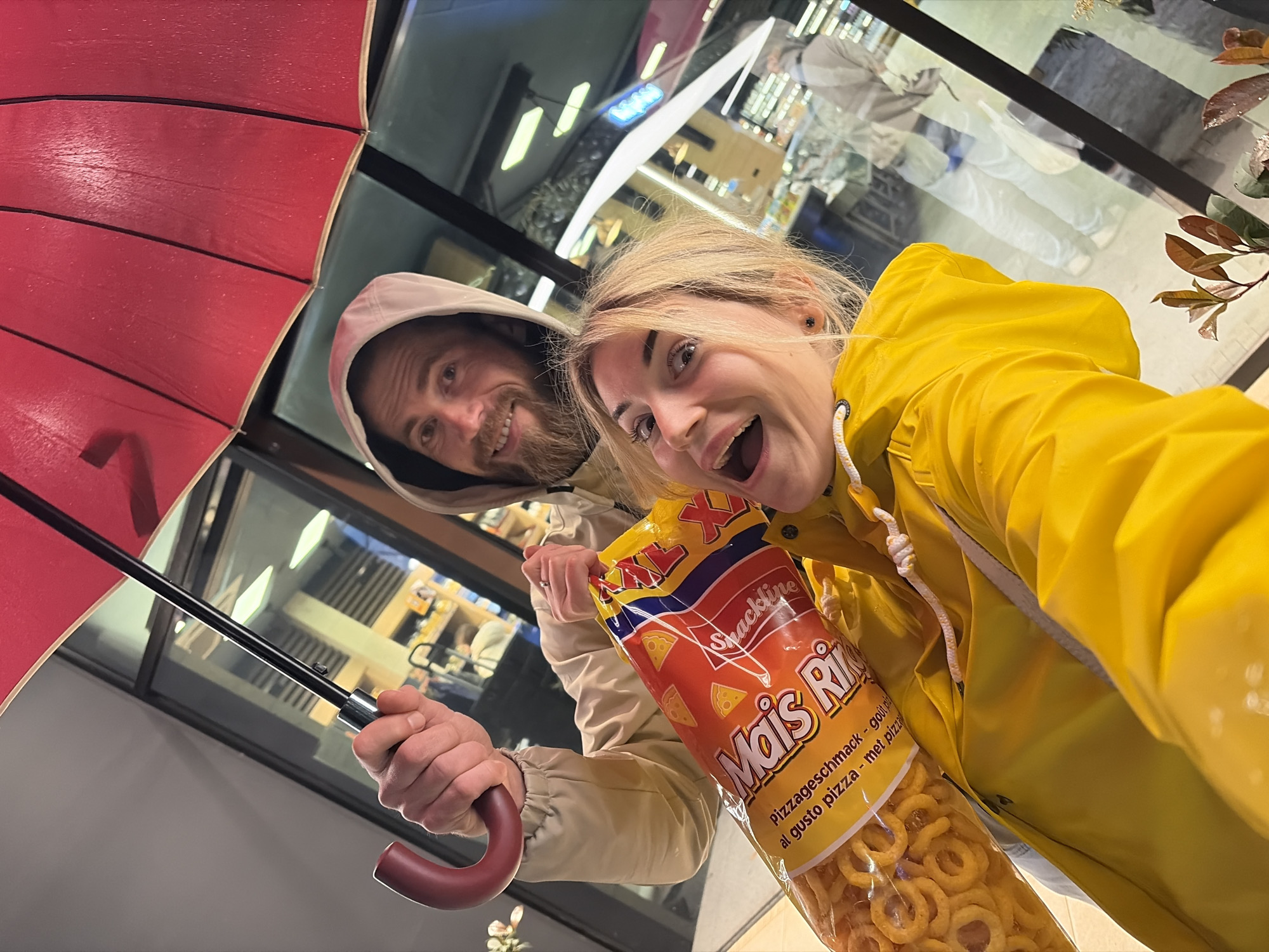 A selfie-style image depicts two individuals, one male and one female, positioned at the entrance of what appears to be a retail establishment in Budva, Montenegro. The background consists of a large glass pane reflecting the interior of a brightly lit store with shelving, fluorescent lighting, and blurred figures of other individuals, suggesting a supermarket or similar public venue.
The male, with a beard and wearing a light-colored hooded jacket, is visible in the upper portion of the frame. He is smiling and holds an open, wet, red umbrella with his right hand. A silver-colored ring is present on the fourth finger of his left hand. He also holds the top of a large, transparent plastic bag of "Snackline Mais Ringo" snacks, marked "XXL" and indicating "Pizza Geschmack" (pizza flavor) in German, Italian, and Dutch.
The female, positioned closer to the camera, has blonde hair and a wide-eyed, open-mouthed expression. She is wearing a bright yellow rain jacket with a white drawstring. She holds the lower section of the same large snack bag. The presence of the wet umbrella and rain jackets suggests recent exposure to rain. The specific address or street location within Budva is not discernible from the visual evidence.