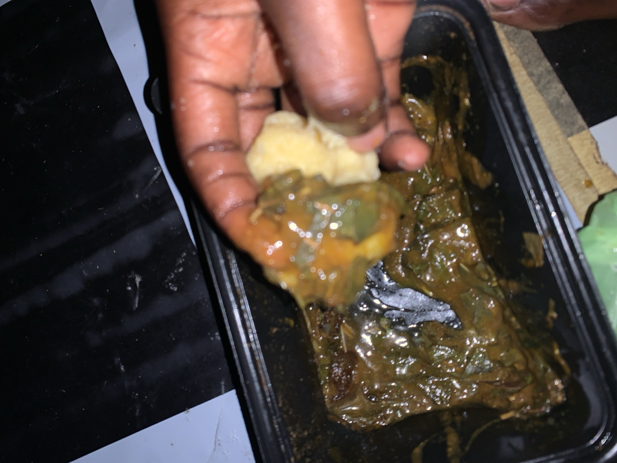 In Rafin Par, Nigeria, a hand is shown holding a piece of food over a black rectangular container filled with a dark green, soupy stew. The stew appears to contain leafy vegetables and possibly some meat or fish. The hand is dark-skinned, and the food being held consists of a light-colored, soft substance, possibly dough or yam, mixed with the stew. The container is placed on a surface that appears to be a table with a black and white checkered pattern. The lighting is dim, and the image is slightly blurry, suggesting it was taken indoors with low light conditions.