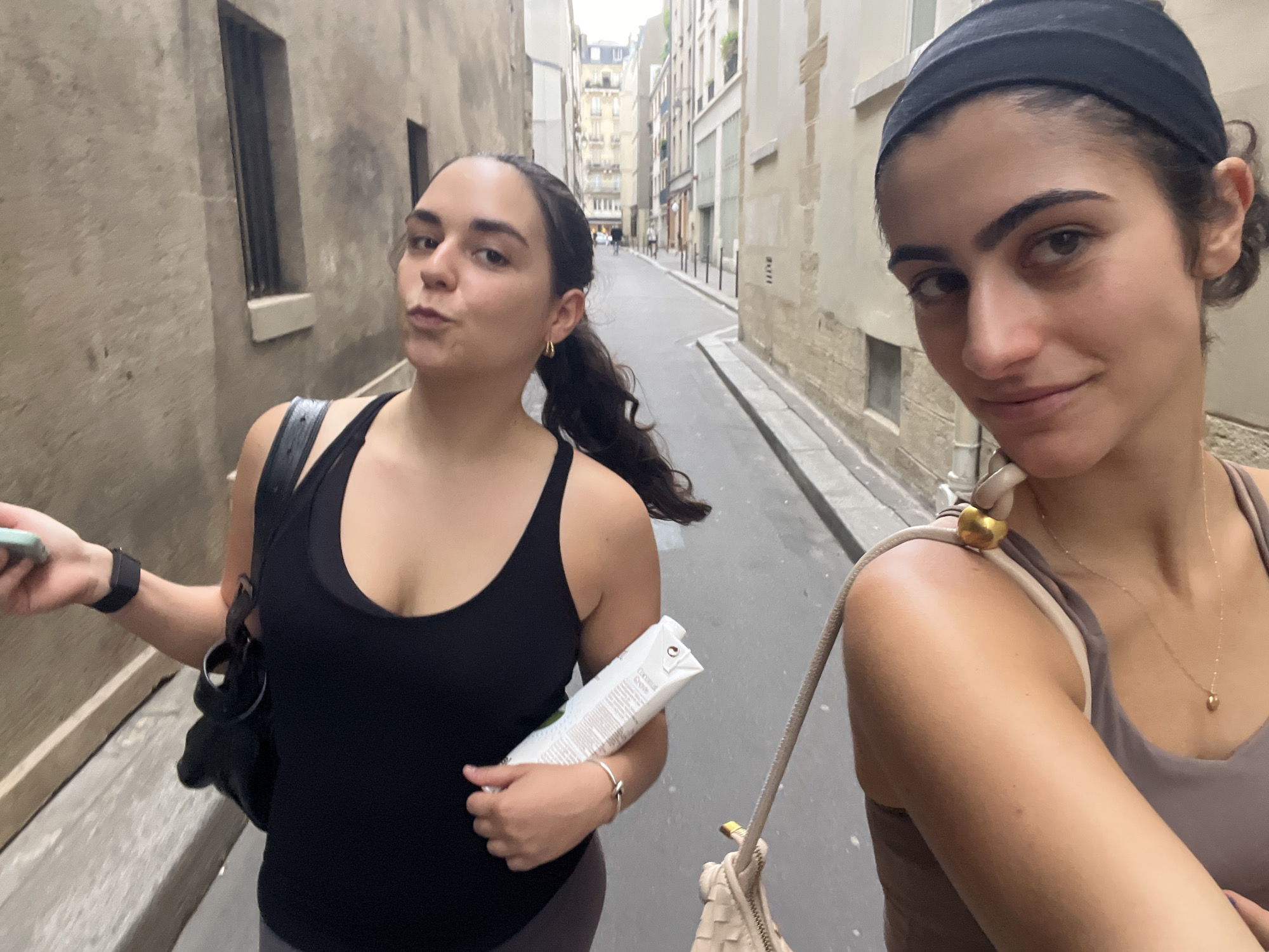 Two women are walking down a narrow, European-style street, possibly in Paris, France. The woman on the left is in the foreground, facing the camera with a playful pout. She has dark hair pulled back in a ponytail and is wearing a black tank top and dark leggings. She holds a white carton in her right hand and her left hand is holding a cell phone. A black bag is slung over her left shoulder. The woman on the right is closer to the camera and is looking to her right, smiling. She has dark hair covered by a black headband and is wearing a taupe athletic top with a thin gold necklace. She has a beige purse with a gold accent strap over her right shoulder. The street is lined with tall, old buildings, suggesting an urban environment. The lighting suggests it is daytime, possibly overcast. The scene appears to be a casual outing, perhaps exploring the city. The carton in the woman's hand has visible text, but it is not legible in the image.