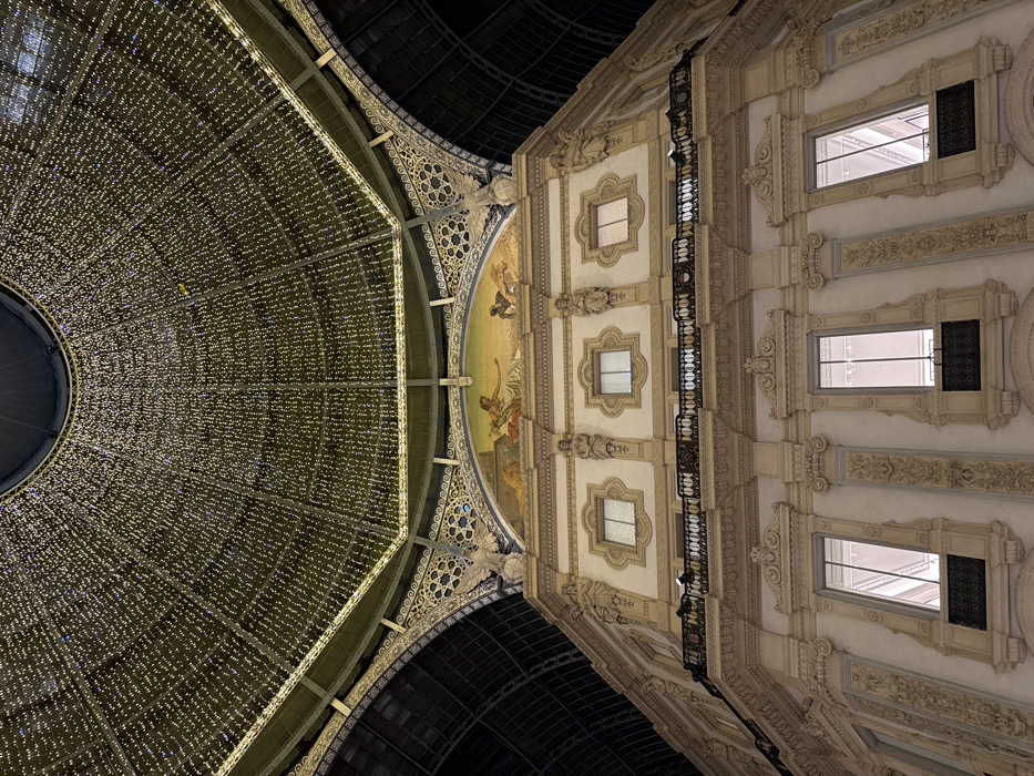 The image displays an upward view within the Galleria Vittorio Emanuele II in Milan, Italy, during nighttime. The upper section is dominated by the expansive, ornate glass and iron dome, which is heavily adorned with thousands of vertically strung, warm-white LED lights, creating a luminous, cascading effect. A dark, circular oculus is visible at the dome's zenith.
Below the dome, a multi-story building facade constructed from light-colored stone or stucco is presented. Directly beneath the dome's elaborate metalwork, a large, semi-circular mural or mosaic depicts classical figures in various poses and attire. To the right of the mural, a decorative sculpture of an eagle is mounted on a pedestal.
The facade below the mural features two levels of classically designed elements. The upper level includes multiple rectangular windows set within decorative frames, flanked by pilasters and sculptural figures. Below this, a prominent black wrought-iron balcony with an ornate railing spans the width of the building. The lowest visible level showcases three tall, rectangular windows with internally illuminated spaces, each framed by elaborate architectural details and featuring small, decorative wrought-iron balconies beneath them. The entire visible structure is characterized by extensive ornamentation, including intricate relief work, cornices, and balustrades. The scene is devoid of people or direct human activity.
