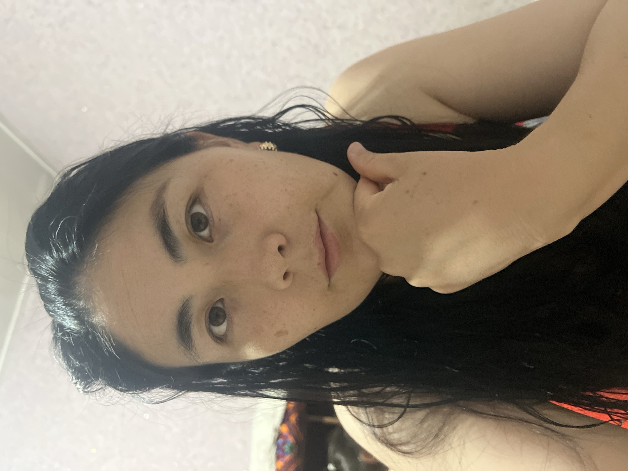 A close-up, selfie-style portrait of a woman in Kara-Dıykan, Kyrgyzstan. The woman has long, dark hair and brown eyes. She is looking directly at the camera with her chin resting on her hand. She is wearing a red top. The background appears to be a light-colored textured wall and a corner of a yurt with colorful fabric.