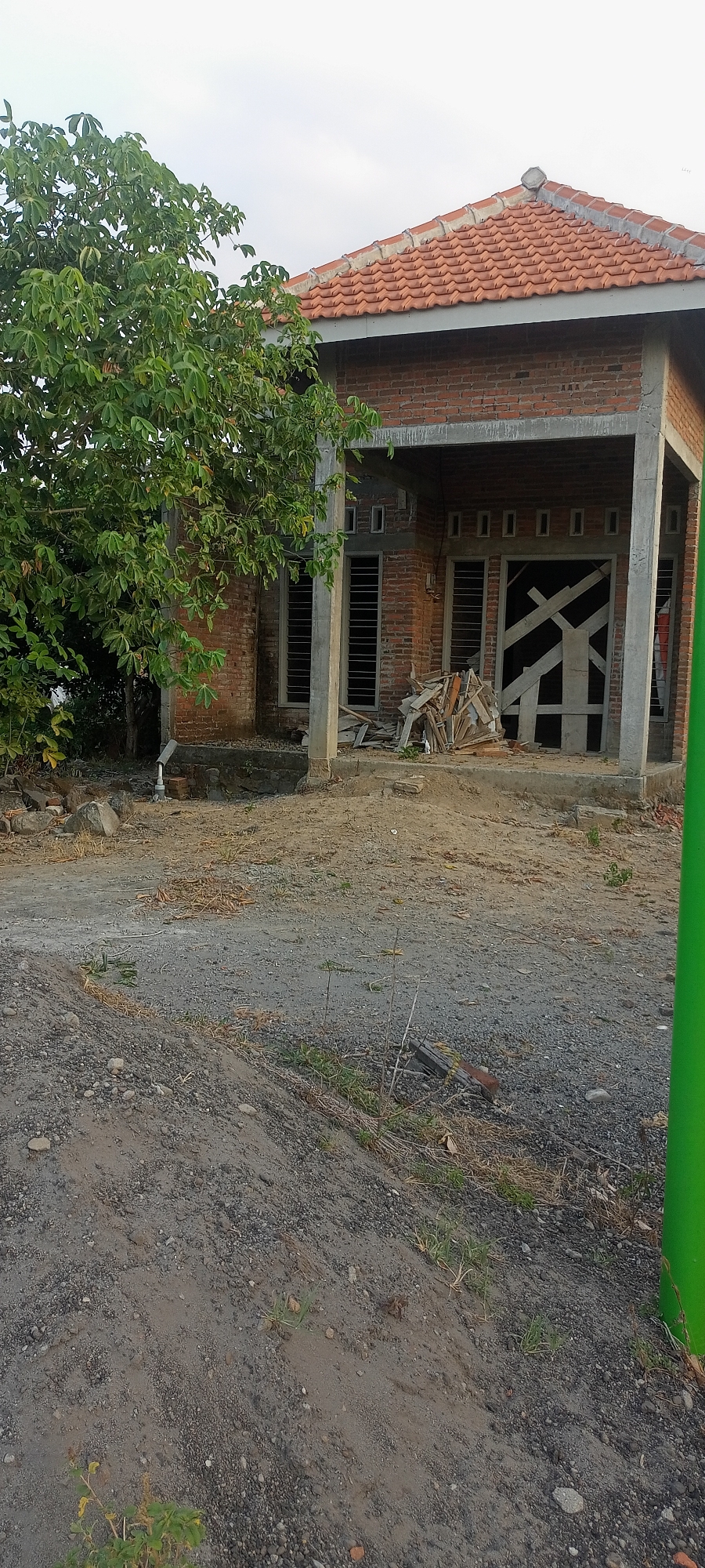 The image shows an exterior view of a house under construction in Gempolklutuk, Indonesia. The house is made of brick with a tiled roof. The front of the house features a covered porch supported by concrete columns. Several windows are visible, some with blinds. The entrance to the house is boarded up with wooden planks. A pile of wood debris is located in front of the entrance. To the left of the house, a large tree with green leaves is present. The ground in front of the house is a mix of dirt, gravel, and scattered rocks. There are no people visible in the image. The sky above is overcast.
