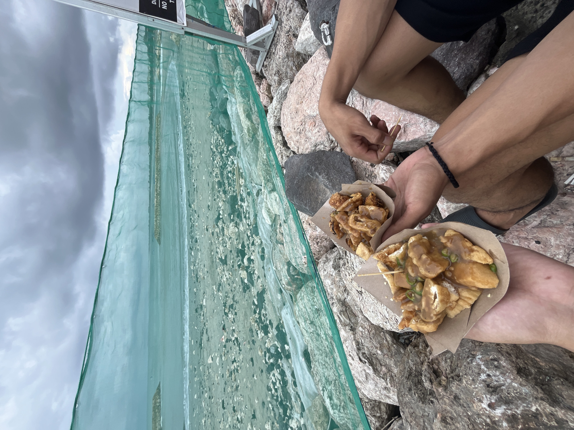 This image captures a casual outdoor scene in Kuta, Indonesia, depicting two individuals enjoying street food. The perspective is a high-angle shot, focusing on the people's lower bodies and the food they hold, set against a rocky background with a distant water body.
Two individuals are present, visible by their legs, arms, and hands. One person wears black shorts and dark sandals, while the other's attire is also casual. One wrist features a braided black bracelet. They appear to be sitting or squatting on rugged, multi-colored rocks that form the ground.
Each person holds a paper cone or bowl containing fried food, prominently featuring irregularly shaped pieces, likely fried tofu (tahu goreng), generously covered in a thick, light brown sauce. Green, thinly sliced chilies are sprinkled on top as a garnish. A wooden toothpick is inserted into one of the servings, indicating it's ready to eat.
In the mid-ground, a light green mesh fence or net stretches horizontally, separating the rocky foreground from the background. Beyond this netting, an expansive area that looks like a sandy or pebble beach transitions into calm water, possibly the sea or a large lake. The sky is overcast with heavy, grey clouds, suggesting a cloudy daytime.
A white sign with partial black text is visible in the upper section of the image, showing fragments such as "DISC", "NO", and "T". The overall impression is one of a relaxed, informal moment spent enjoying local snacks by the water.