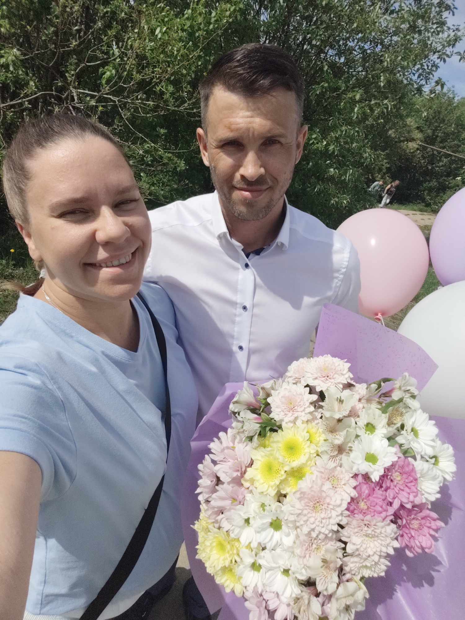 A selfie captures a man and a woman outdoors, holding a bouquet of flowers and balloons. The woman, positioned on the left, wears a light blue t-shirt and a black strap. She is smiling. The man, on the right, wears a white button-down shirt. He also appears to be smiling. They are in front of several pastel-colored balloons, and holding a bouquet that includes white, pink, and yellow chrysanthemums, wrapped in purple paper. The background features green foliage and a partially visible figure in the distance. The location is Gremyachinsk, Russia.