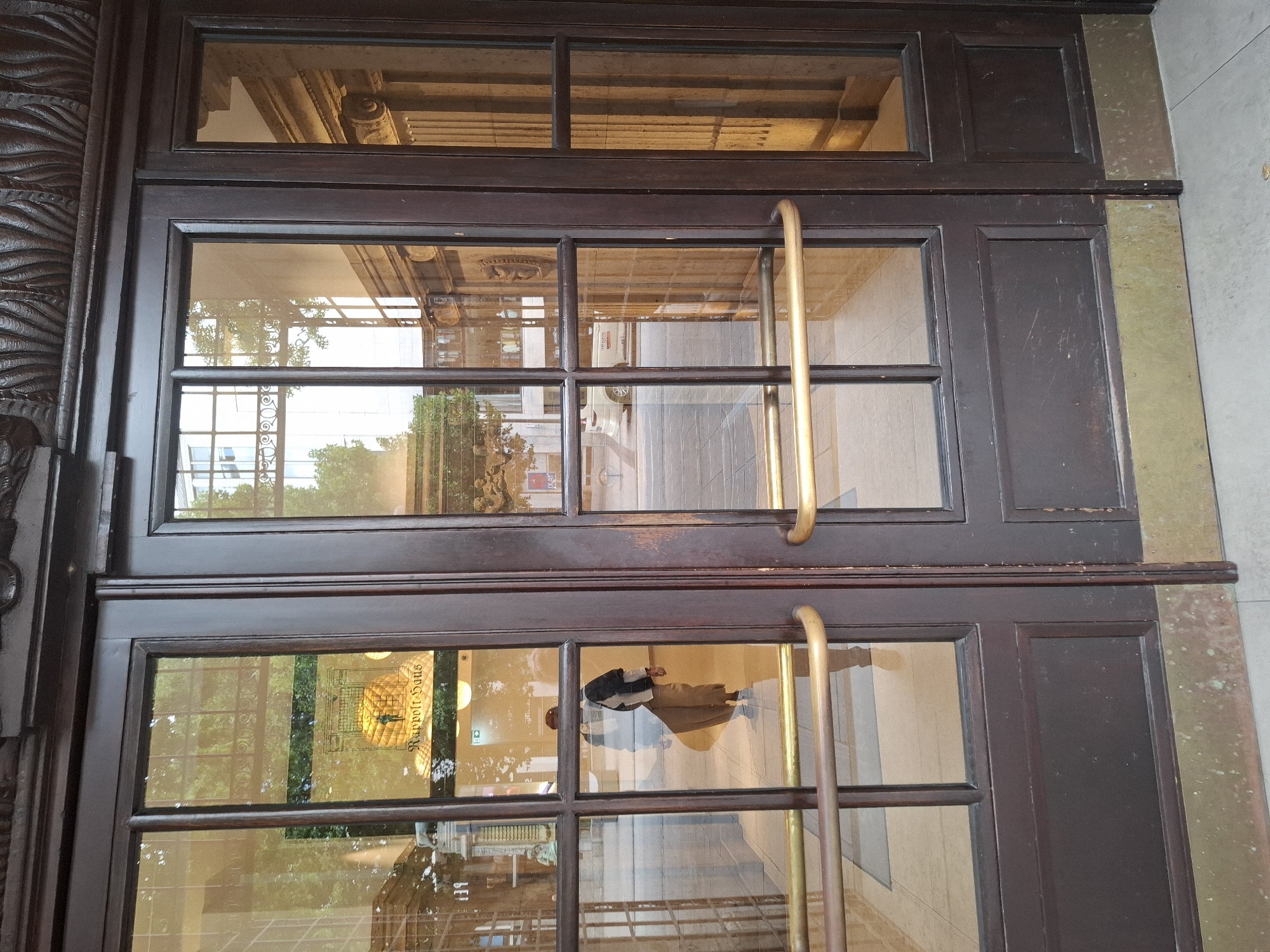 The image shows a close-up of large, dark wooden double doors with glass panels. The doors have ornate carvings at the top. There are brass door handles. The glass reflects the environment outside and inside. In the reflection of the left door, a person is visible from the waist down, wearing light-colored pants and dark shoes. Above this, a sign is visible with text that appears to read "Rappolt-Haus". The scene appears to be in a city, with buildings and trees reflected in the glass. The location is identified as Hamburg, Germany.