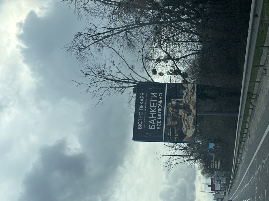 The image shows a billboard on the side of a highway in Chabany, Ukraine (latitude: 50.339, longitude: 30.412). The billboard advertises a restaurant called "Bistro Pekary" with the words "Banketi Vse Vkljucheno" written in large, white letters, which translates to "Banquets All Inclusive" in English. The billboard features a picture of a table set with a buffet of food.  The billboard is positioned on a metal pole on the side of the highway, and there are trees in the background. The road is wet, and the sky is overcast. The road is empty except for a few cars in the distance.  The image was likely taken from a vehicle on the highway.