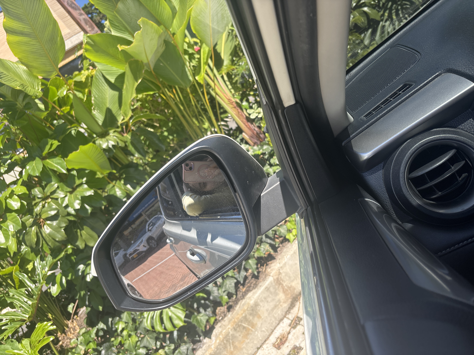 The image shows a car's side mirror from the interior perspective. The mirror reflects another car parked behind, with a brick-paved area visible. Lush green foliage is abundant outside the car window, including large leaves and smaller plants. Inside the car, parts of the dashboard, air vent, and door frame are visible. In the side mirror's reflection, a person is discernible holding a smartphone, likely the photographer. Based on the location tag, the photo was taken in Llanogrande, Colombia.