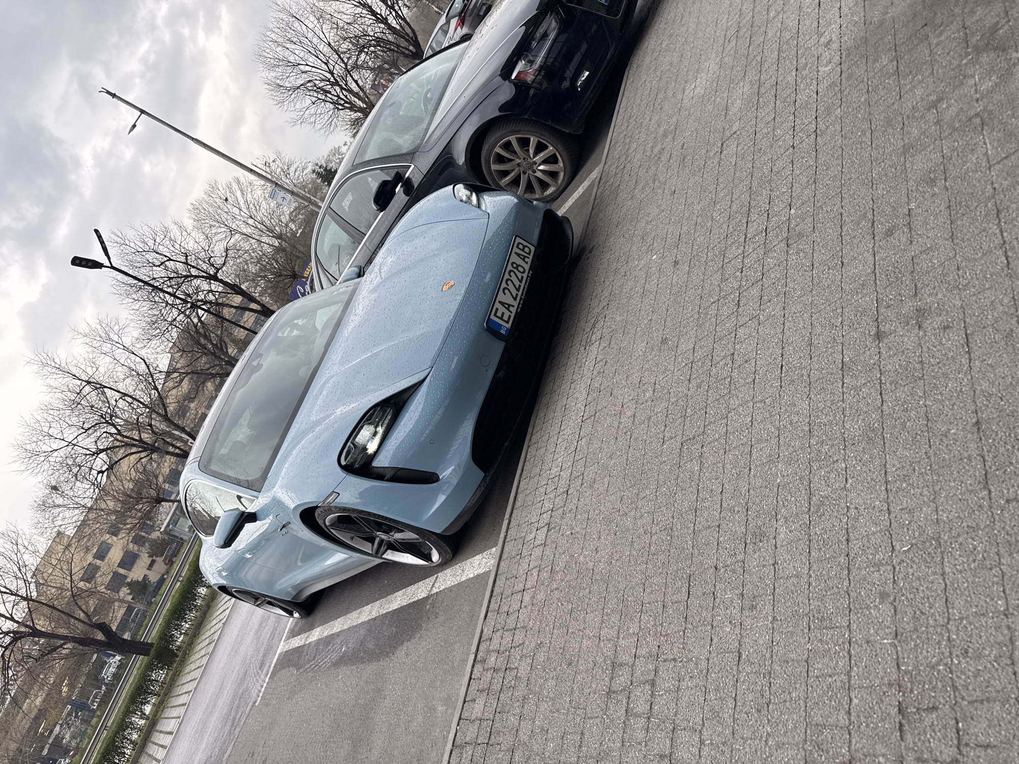 The scene depicts an outdoor parking area in Plovdiv, Bulgaria, identified by the "EA" prefix on the vehicle's license plate. A light blue Porsche Taycan electric vehicle, covered in water droplets from precipitation, is parked in the foreground, facing forward and slightly angled towards the right. Its front headlights are illuminated. The Bulgarian license plate "EA 2228 AB" is affixed to the front bumper. To the right of the Porsche, a dark-colored Audi sedan is partially visible, also appearing wet. Both vehicles are situated on asphalt pavement marked with white parking lines. The immediate foreground shows a section of gray rectangular paving stones. The background features several bare deciduous trees under an overcast sky. A multi-story building with light-colored walls and dark windows is visible behind the trees. Two streetlights are present, one modern and one older, along with a directional sign. No individuals or active interactions are present in the image; the scene is static.
