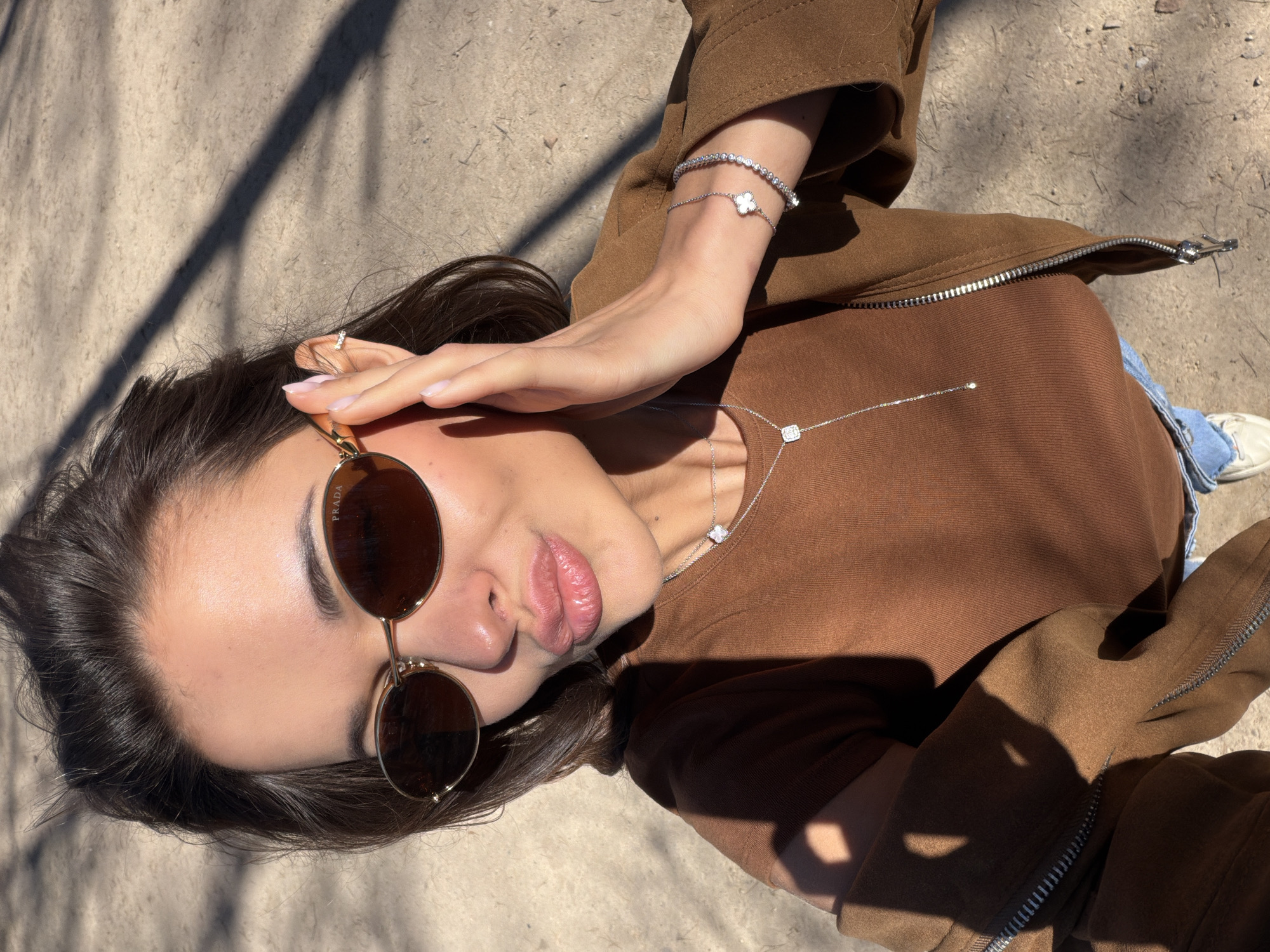 The image shows a young woman lying down on an outdoor surface, angled such that she is looking up towards the camera. Her dark hair frames her face, and she is wearing round, brown-tinted sunglasses. Her left hand is raised, with fingers touching the top edge of her sunglasses, revealing light pink nail polish. Her lips are slightly pursed.
She is dressed in a brown long-sleeved top and a brown zippered jacket. A silver-toned zipper is visible on the jacket. Her attire is complemented by blue jeans and white sneakers, partially visible on her lower right side.
Jewelry worn includes two silver-toned bracelets on her right wrist: one is a tennis bracelet with clear stones, and the other is a delicate chain bracelet featuring a white clover-shaped charm. Around her neck, she wears two silver-toned necklaces: one with a white clover charm and another longer chain with a small square pendant containing a clear stone. A small, sparkling hoop earring adorns her left ear.
Strong sunlight creates distinct, elongated shadows across the textured, light brown ground and parts of her clothing, indicating a sunny daytime setting. The ground appears to be dirt or a coarse outdoor path. The visible text "PRADA" can be seen on the right arm of her sunglasses. The overall scene suggests a casual, stylish moment captured outdoors in Krasnoyarsk, Russia.