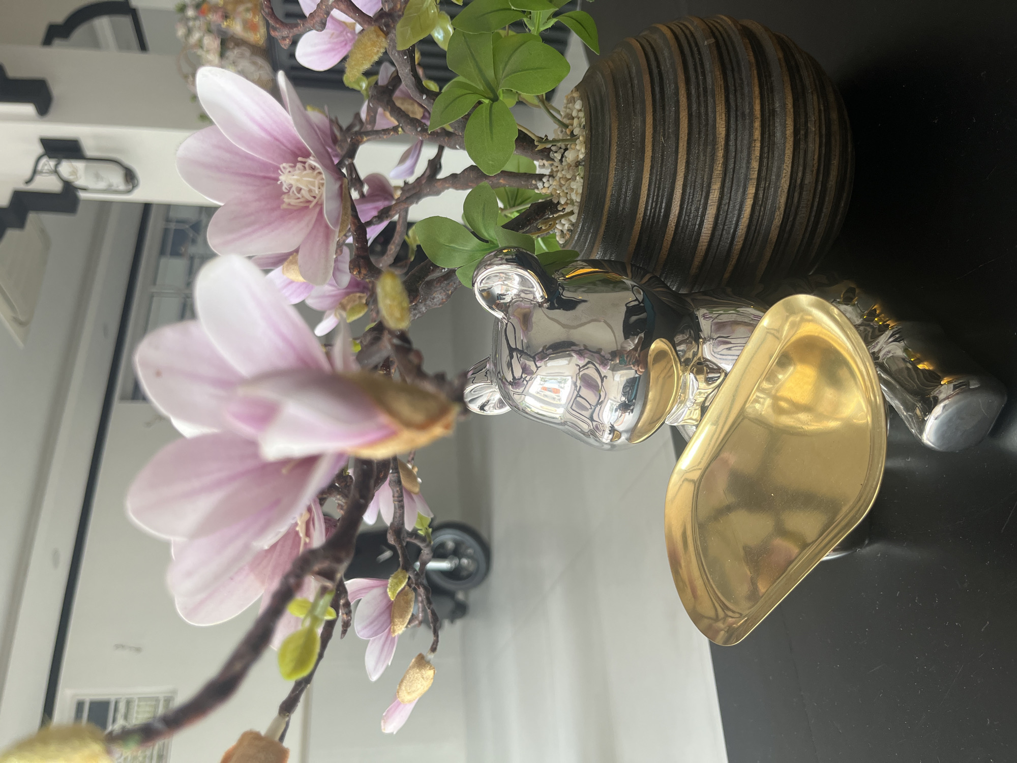 The image shows a close-up of a decorative arrangement on a dark surface. In the foreground, a shiny, silver, bear-shaped figurine is positioned next to a golden tray. Behind the figurine and tray, a potted plant with green leaves is housed in a dark, textured pot. The plant appears to be part of a larger decorative arrangement that includes branches with pink magnolia-like flowers. In the background, out of focus, a light-colored tiled floor is visible, and a black scooter with a single wheel is parked. The overall setting seems to be an indoor space, possibly a lobby or a commercial area, with a modern aesthetic. The lighting suggests an indoor environment with diffused natural light.