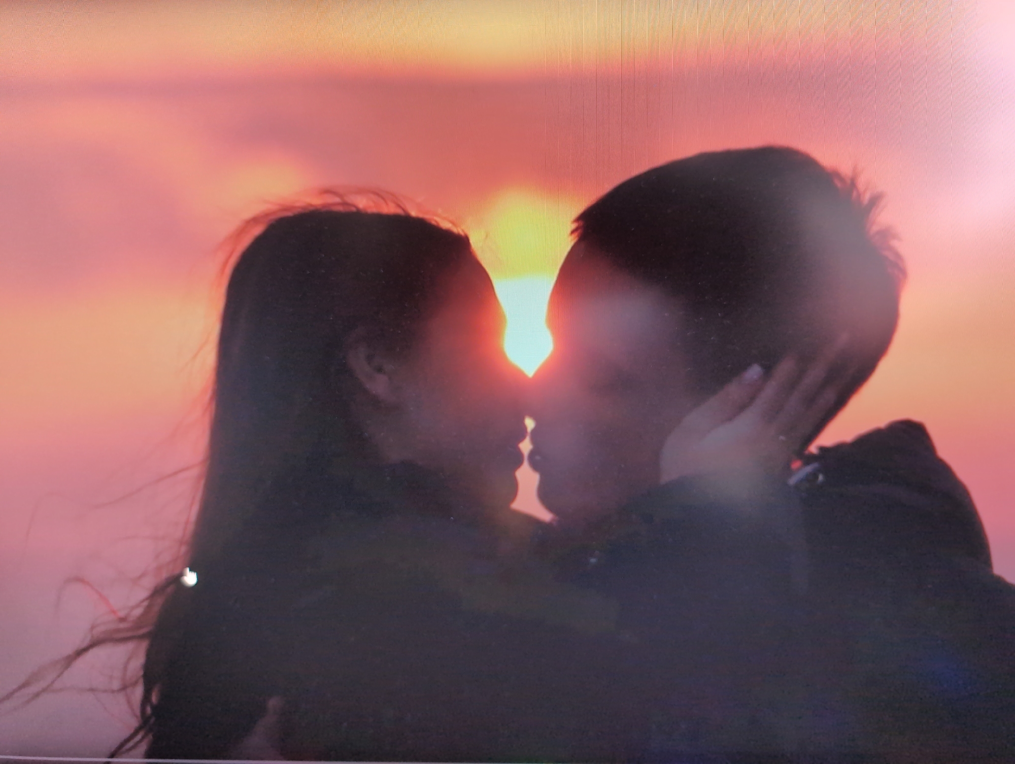 A silhouette of two people kissing is visible against a backdrop of a sunset in shades of orange and pink. The person on the left has long hair, while the person on the right has shorter hair and is holding the other person's face with their hand. The location is not identifiable as Izhevsk, Russia from this image.