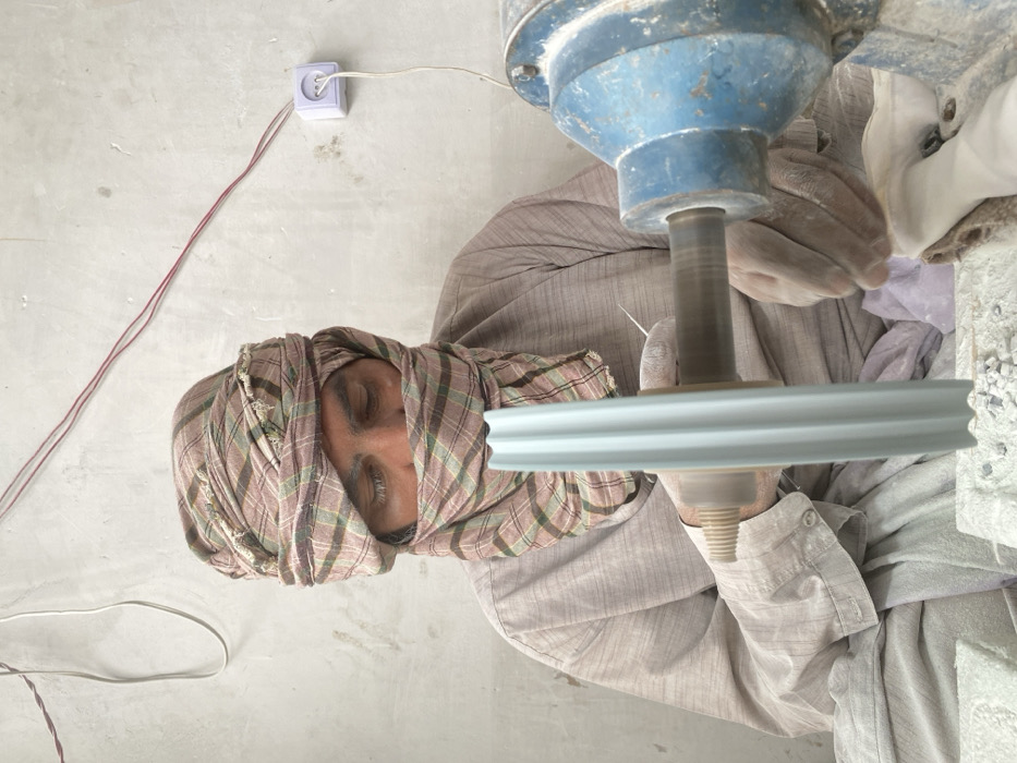 In a workshop in Kandahar, Afghanistan, an adult male artisan is seated, engaged in a stone carving or polishing operation. He wears a light-colored, long-sleeved garment and a plaid headwrap that covers his head and lower face, with his eyes partially visible. His hands, appearing calloused and dusted with fine white particulate matter, manipulate a light blue, vertically fluted object against a rapidly spinning wooden mandrel. This mandrel is powered by a blue industrial electric motor, partially visible on the right side of the frame. The immediate work surface in the foreground is covered with white dust and small, irregular stone fragments. The background consists of a plain, light-colored wall exhibiting some discoloration. Exposed red and white electrical wires run across this wall, connecting to a white, surface-mounted European-style power outlet. A white electrical cord extends from this outlet, presumably powering the motor.