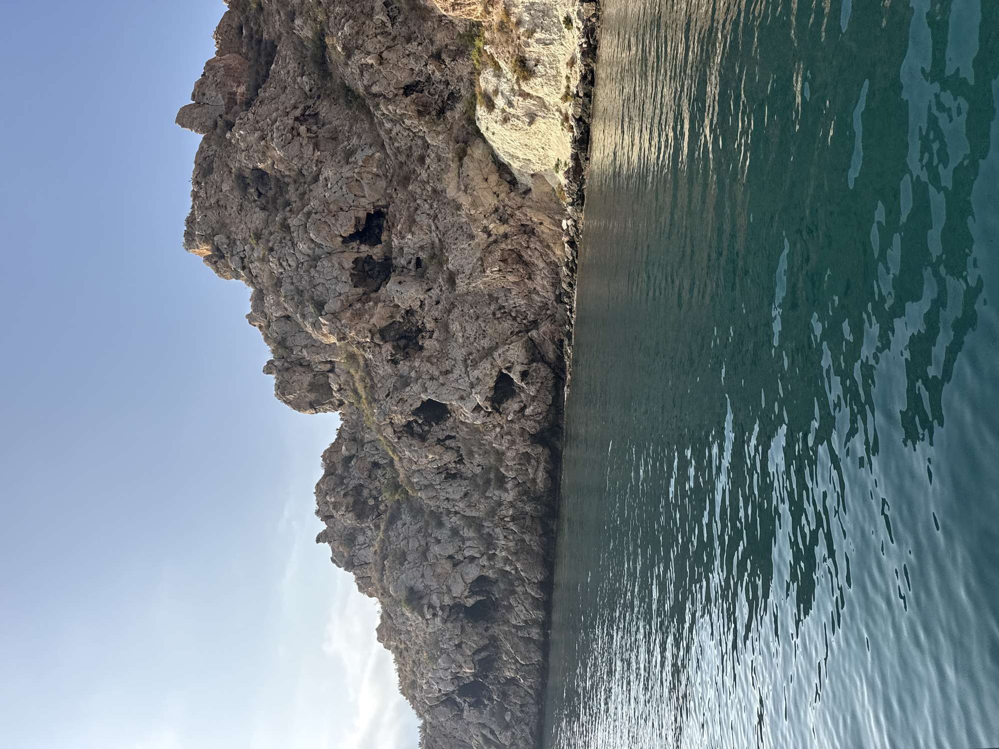 The image captures a coastal scene, likely in or near Águilas, Spain, featuring a rugged, rocky cliff face rising from the sea. The cliff is characterized by its uneven texture, numerous small caves, and sparse vegetation. The rock formations display a palette of earthy tones, ranging from light tan to darker browns and grays. The sea in the foreground is a deep, tranquil teal, with subtle ripples reflecting the light from the sky. The sky above is a clear, pale blue, with a faint haze near the horizon, suggesting either early morning or late afternoon light. The overall impression is one of natural beauty and untouched landscape.