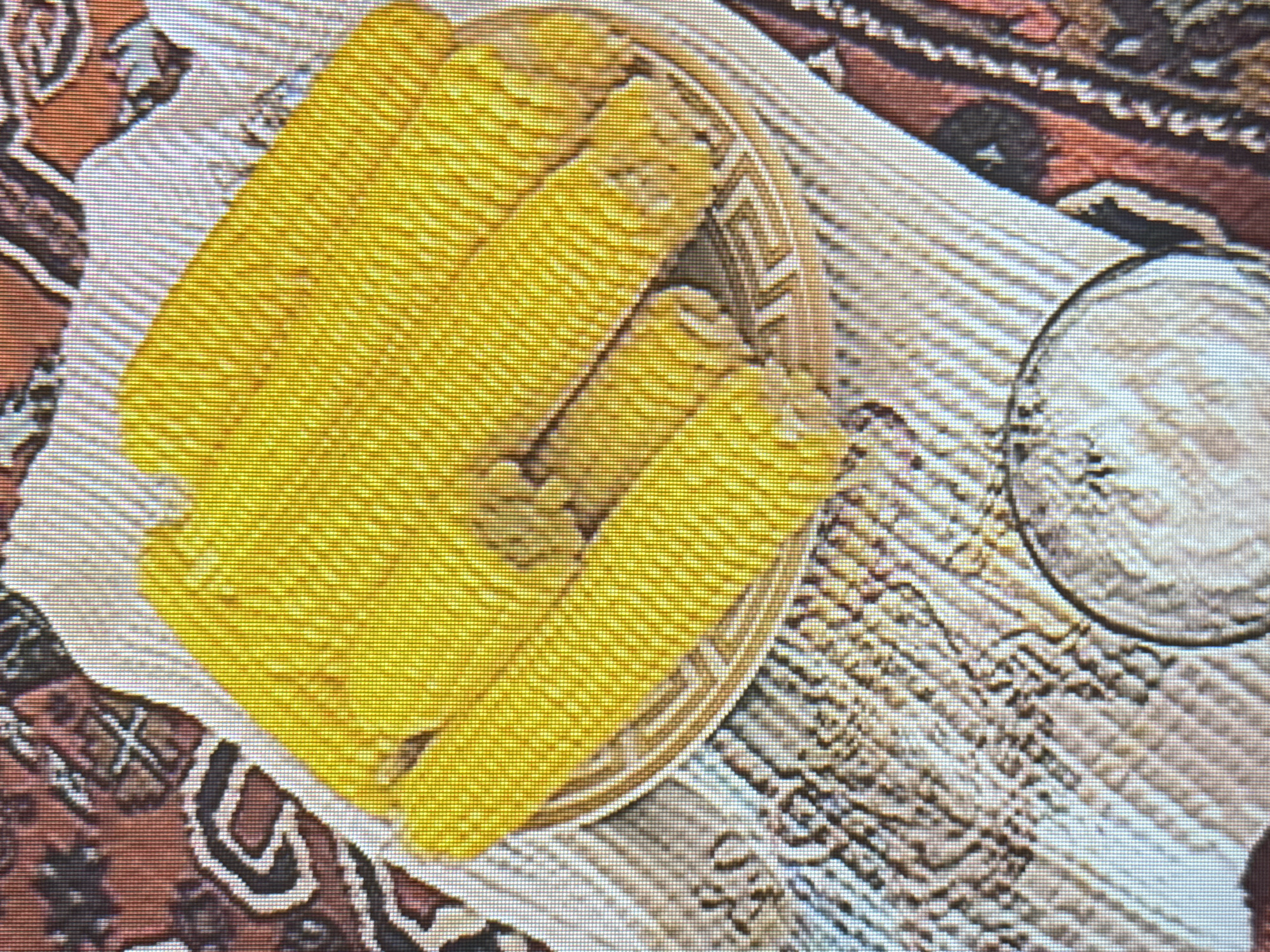 The image shows several cobs of corn arranged in a woven basket. The corn is bright yellow and appears to be cooked. The basket is placed on a white cloth with a patterned border. In the background, there is a richly patterned rug with red, brown, and white colors. To the right of the basket, there is a glass of what appears to be a light-colored liquid, possibly water or a beverage, and next to it, a small pile of salt or sugar. The lighting suggests it might be daytime, possibly indoors or in a shaded outdoor area. The scene evokes a feeling of a simple, rustic meal or a food preparation setting. The patterned rug suggests a cultural context, possibly related to Armenian traditions given the location context of Yerevan, Armenia. There is no visible text in the image.