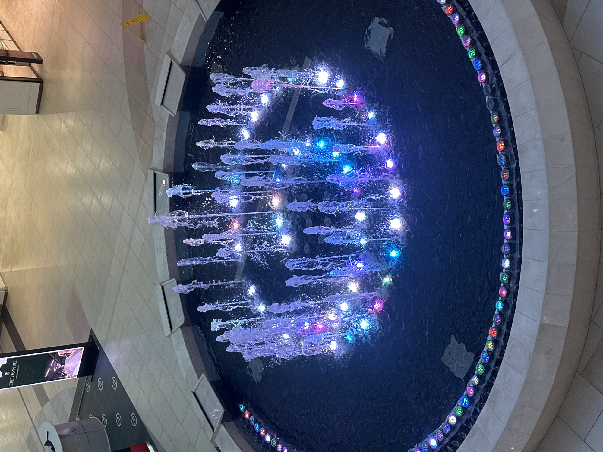 This overhead image depicts a vibrant, circular fountain with illuminated water jets located within a spacious indoor environment, likely a shopping mall or large public building in Sochi, Russia. The fountain features dark water and numerous streams that spray upwards, each lit by dynamic, colorful LEDs in shades of blue, purple, pink, green, red, and white. Additional colored lights are embedded around the rim of the fountain, reflecting off the water's surface.
The fountain is set into a light-colored tiled floor, which shows a grid pattern. Portions of the floor feature darker grey/black tiles with repeating circular logos. To the bottom left of the frame, a tall, vertical digital screen or banner displays a logo and the Cyrillic text "СВЕТСКИЙ ЛЕС" (Svetsky Les). Below this, a cylindrical object also appears to have the same text. In the upper left corner, a yellow "wet floor" warning sign is partially visible on the tiled ground.
The overall scene suggests an artificial, well-lit environment designed for public gathering or commerce. The active water display and shifting colorful lights create a dynamic and appealing focal point, characteristic of modern urban interiors. The image is taken from an elevated perspective, looking down at the fountain and its immediate surroundings.