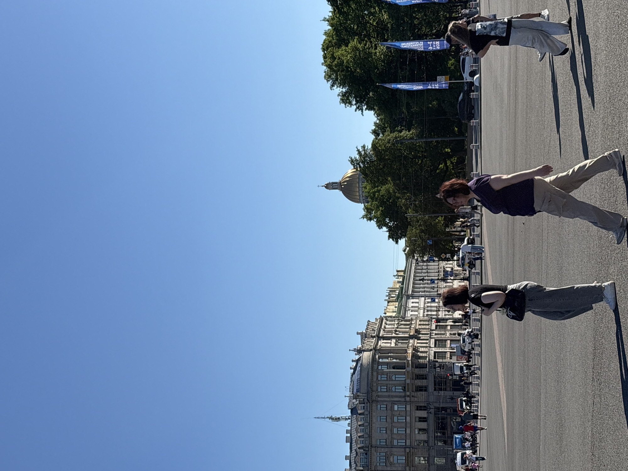 This image captures a bright, sunny daytime scene in central Saint Petersburg, Russia. The golden dome of Saint Isaac's Cathedral is prominently visible in the upper right background, rising above a dense line of green trees. To the left, a row of multi-story historical buildings with ornate, classical architecture lines what appears to be a wide street or square.
The sky is a clear, uninterrupted expanse of bright blue, occupying the upper half of the frame. The foreground and midground consist of a vast, grey paved surface, indicative of a large urban square or thoroughfare, with strong sunlight casting distinct shadows of people and objects.
Three women are walking across the paved area from left to right. The first woman, on the left, has dark hair, wears a black short-sleeved top, light grey wide-leg trousers, and white sneakers, carrying a dark shoulder bag. She is looking to her right. In the center, a woman with reddish-brown hair wears a dark purple short-sleeved top, light khaki trousers, and light-colored sneakers, looking forward. The third woman, on the right, has long dark hair, wears a black short-sleeved t-shirt with a white graphic design, light grey wide-leg trousers, and dark flat shoes, with her hands behind her back and looking slightly to her right. The lower legs and shadow of a fourth person are visible behind her.
In the background, below the historical buildings on the left, several cars, vans, and pedestrians are visible. Near the trees on the right, several tall blue flags with white Russian text are interspersed.