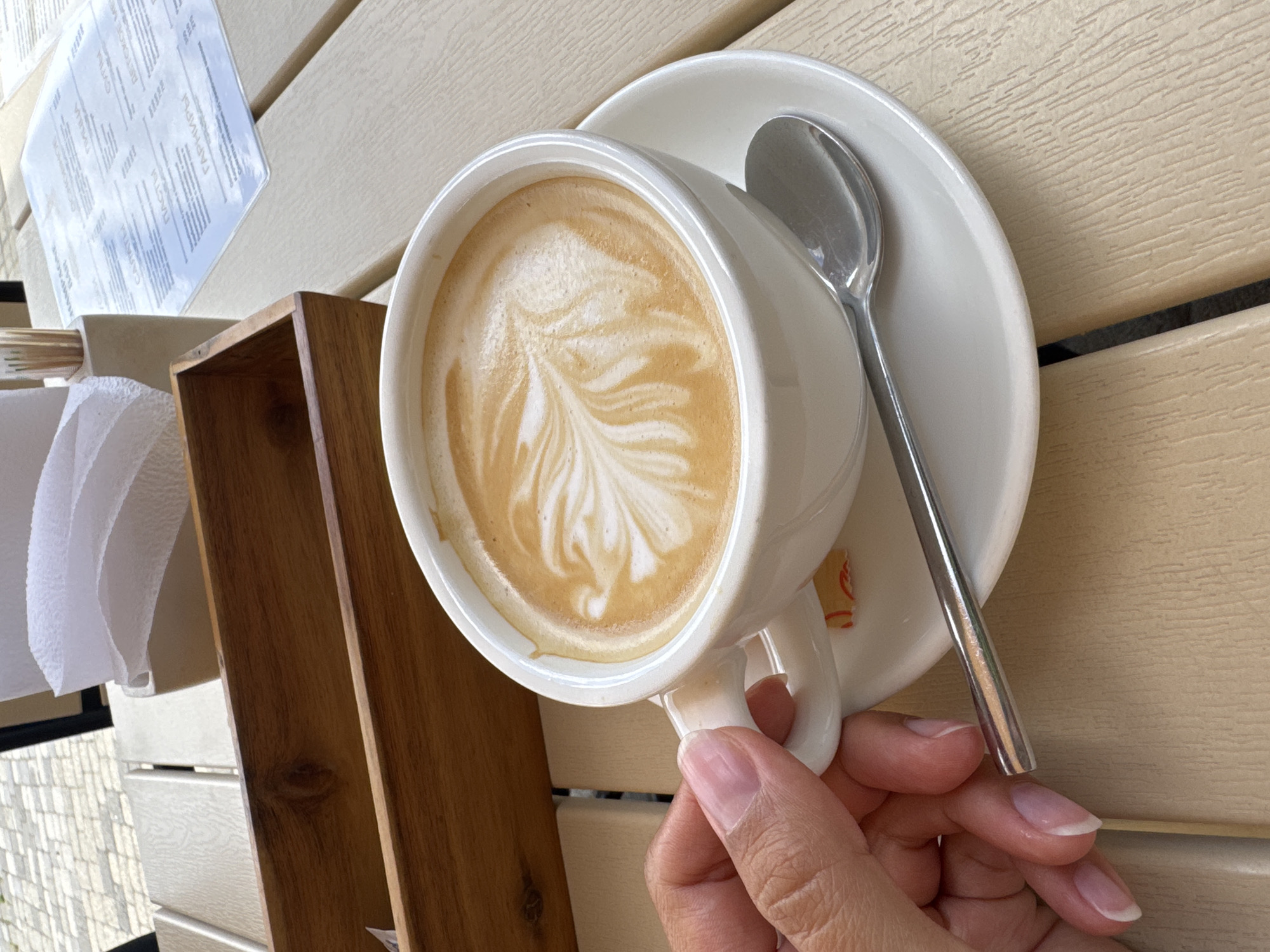 A close-up shot shows a hand holding a white ceramic mug filled with latte art, likely on a wooden table outdoors. The latte art features a fern-like pattern. A silver spoon rests on the saucer. In the background, a wooden holder contains napkins and a pack of toothpicks. A menu is visible on a transparent overlay in the far background. The overall setting suggests a cafe or restaurant environment in Abrau-Dyurso, Russia, given the menu's Cyrillic script and typical cafe offerings.