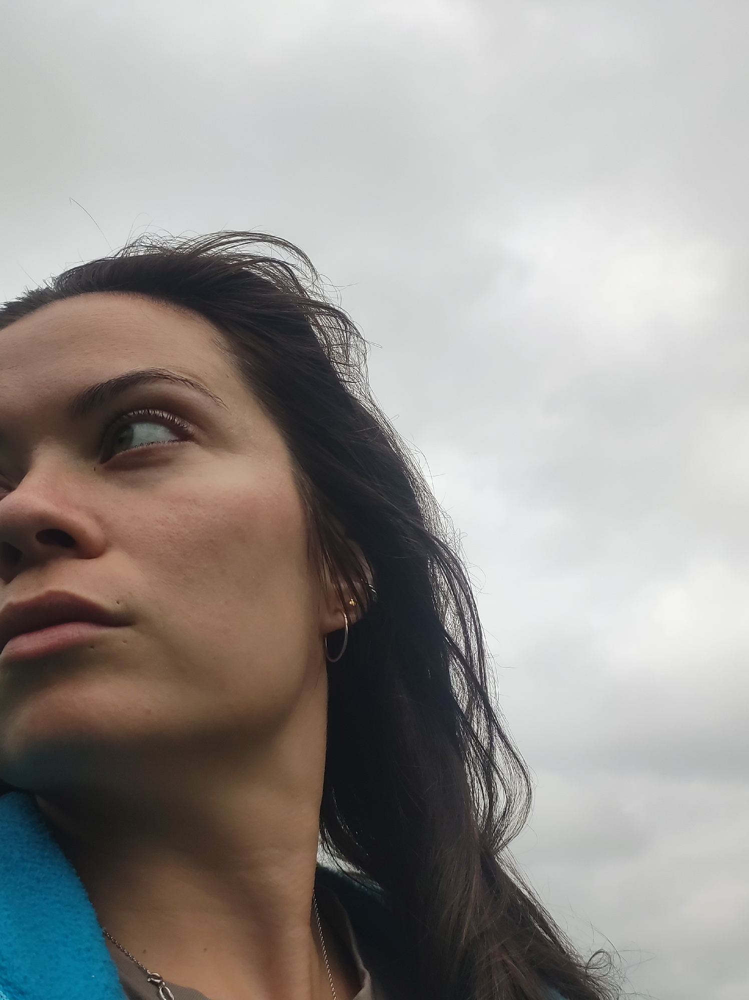 A low-angle medium shot captures a close-up of a young woman's face as she looks upwards and to the left, against a background of an overcast sky. Her dark brown hair, slightly windswept, frames her face. She has dark eyebrows, green eyes with a subtle sparkle, and fair skin with visible pores and a faint blush on her cheek. A small, circular silver earring adorns her right ear. She is wearing a teal-colored jacket and a simple silver necklace. The composition is dominated by her profile and the texture of the sky. There are no discernible elements indicating a specific urban location within the city of Фоки, Russia. The scene appears to be outdoors, possibly in an open area given the sky as the sole backdrop.