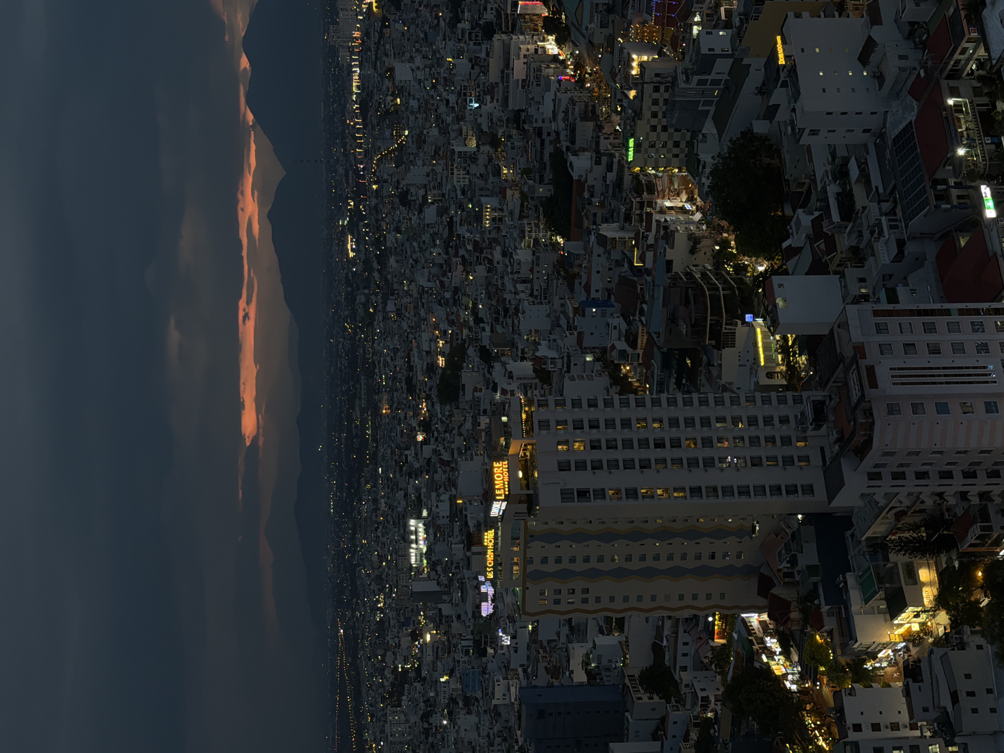 The image shows an aerial view of Nha Trang Ward, Vietnam, at night. The city is densely packed with buildings of varying heights and architectural styles, illuminated by streetlights and the lights emanating from buildings. A tall hotel with visible signage is a prominent feature in the cityscape. The sky is dark, with a hint of color at the horizon where the sky meets the mountains in the background.