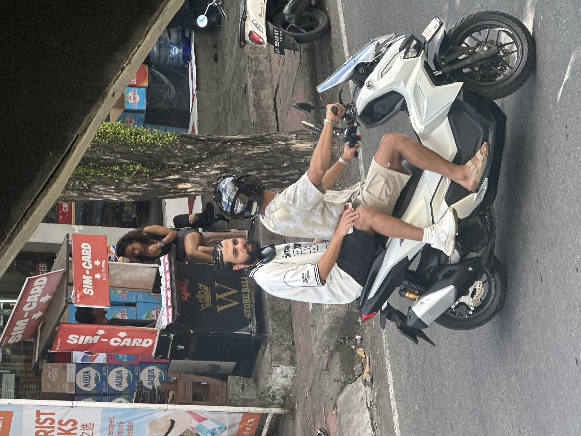 Two men are riding on a white scooter in Ubud District, Indonesia. The man in front is wearing a black helmet and a tan t-shirt, with tan shorts. The man behind him is wearing a white t-shirt with black lettering and black shorts. Both men are wearing casual footwear. The scooter has a transparent windshield and a license plate reading "DK 5540 SY 12 26". The scooter is driving on a paved road with some debris. In the background, there is a street stall selling SIM cards, with several boxes of bottled water and other goods. A woman is sitting at the stall. Trees and other urban elements are visible in the background.