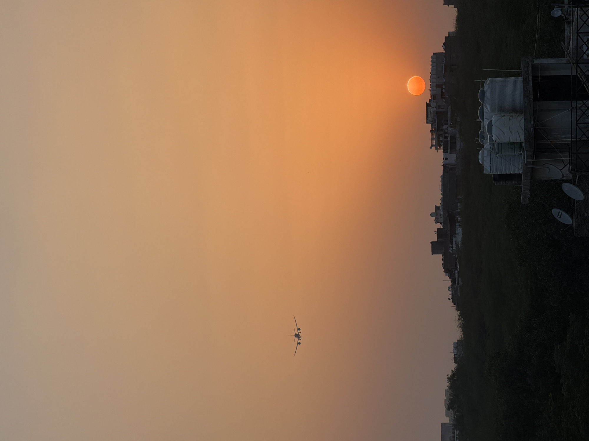 The image depicts a silhouetted airplane flying across a vibrant orange sunset sky over the city of Delhi, India. The sun, a bright orange orb, is partially obscured by a building on the right side of the frame. In the foreground, a rooftop structure with several white water tanks is visible, along with satellite dishes. The lower portion of the image shows a dense area of dark green foliage, suggesting a park or wooded area within the urban landscape. The overall scene captures a moment of transition from day to evening in a bustling Indian metropolis.