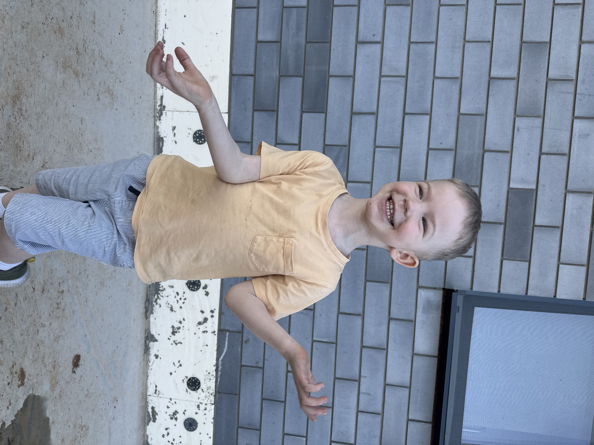 A young boy with light-colored hair is smiling and standing on a concrete and tiled surface. He is wearing a light yellow t-shirt with a pocket and horizontally striped shorts. His arms are raised with his hands open. The background consists of grey brick tiles and a concrete slab with white tiles interspersed with black circular markings. A screen door or window is visible on the right side of the image. The location is Прикамский, Russia.