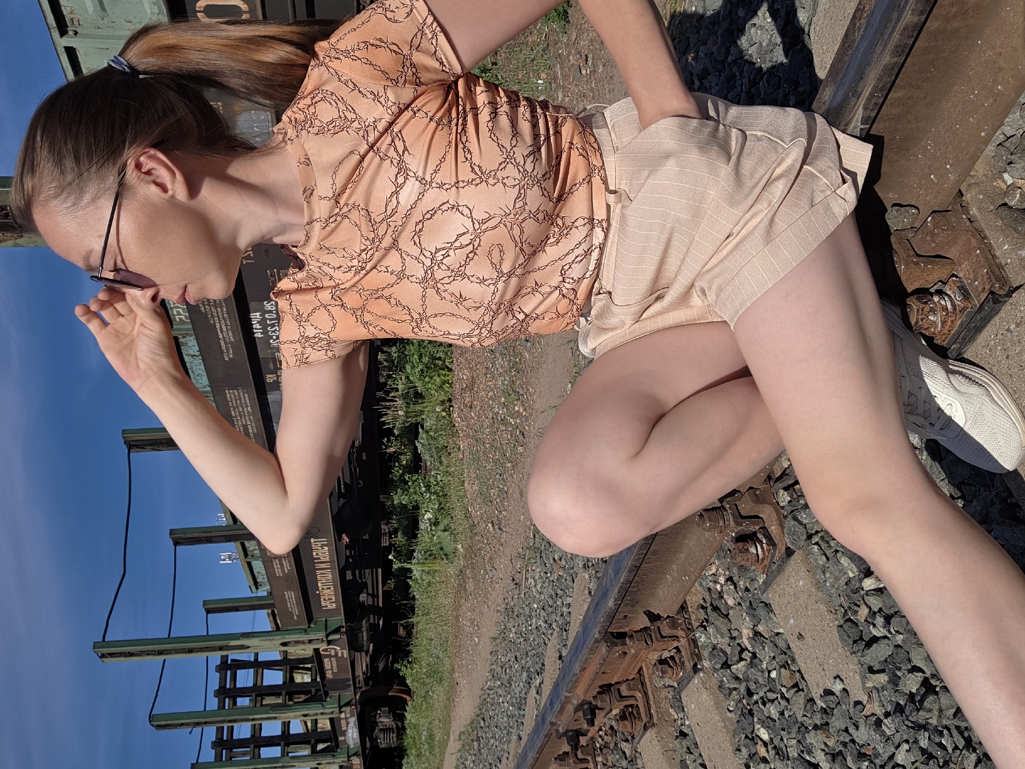 A woman is crouching on a railroad track next to a freight car in Chaikovsky, Russia. The woman has fair skin, dark hair pulled back in a ponytail, and is wearing sunglasses, a peach-colored short-sleeved shirt with a dark brown barbed wire design, peach-colored shorts with white stripes, and white sneakers. Her left hand is raised near her face and her right hand is in her shorts pocket. The freight car is dark green with some rust, and Cyrillic text is visible. The railroad tracks are rusty and surrounded by gray gravel. Patches of green grass are visible in the background. The sky is blue.