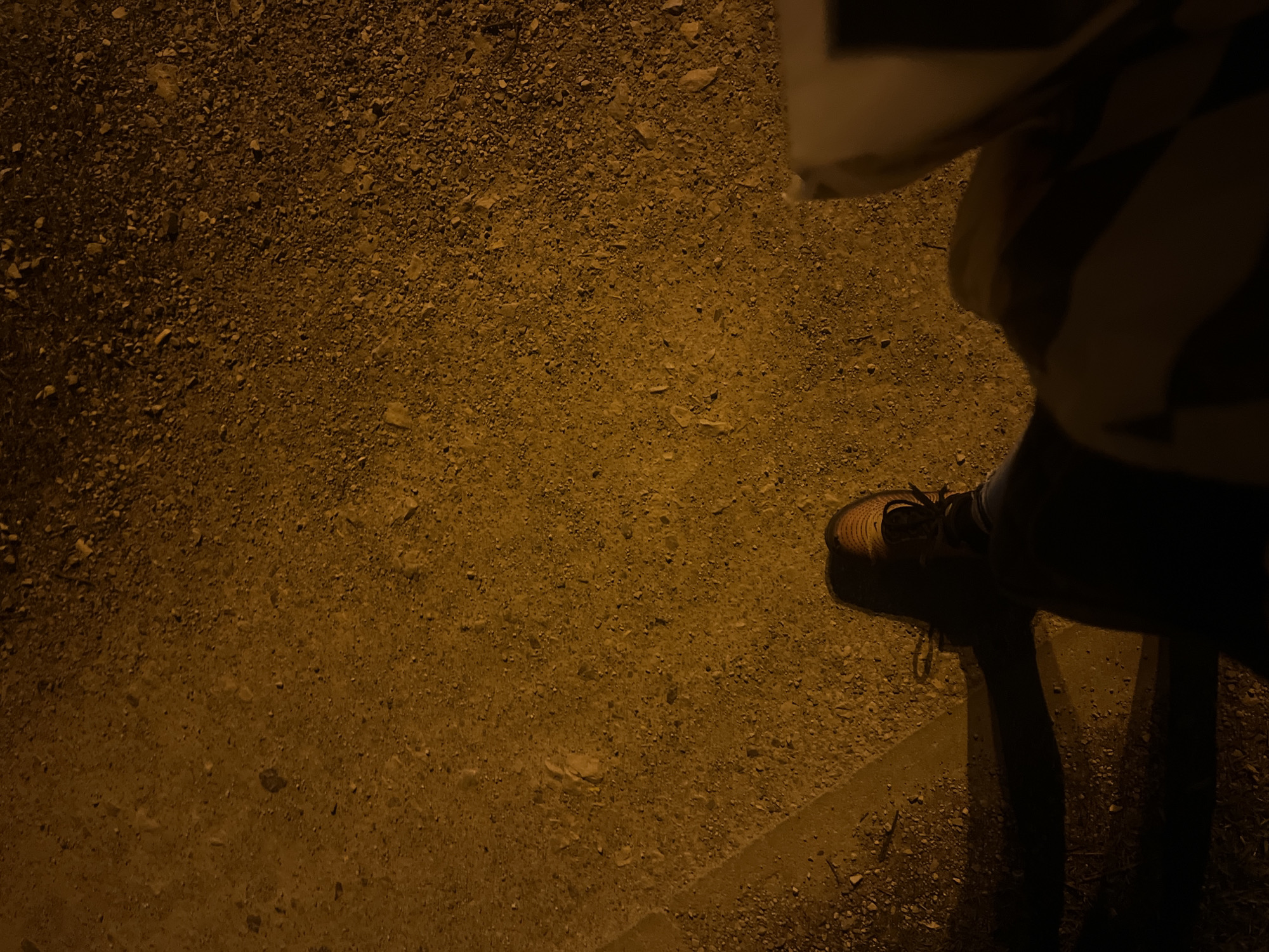The image shows a person standing on a gravel surface next to a sidewalk. The person is wearing a tan shoe. The lighting suggests it is nighttime or a dimly lit area. The location is unknown, it cannot be confirmed whether it's Sofia, Bulgaria.