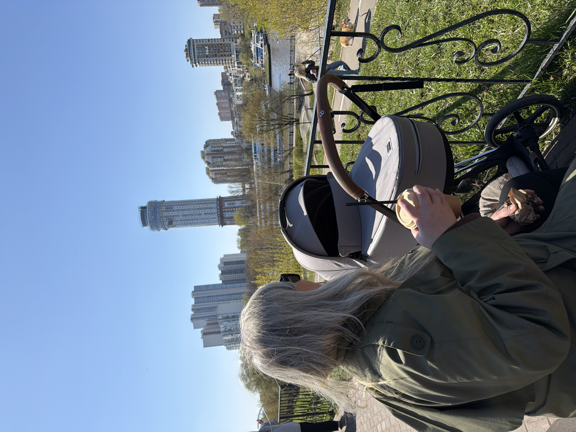 A woman with long grey hair, wearing sunglasses and an olive-green jacket, is sitting on a bench beside a grey pram. She is holding a cup in one hand and a crumpled paper bag in the other. A wrought-iron fence separates her from a grassy area with a pathway, where a dog is walking. In the background is a river and several high-rise buildings, indicative of an urban landscape. This view is likely in Khimki, Russia, based on the architecture and geographical elements. The sky is clear and blue. The photograph is taken from slightly above and behind the woman.