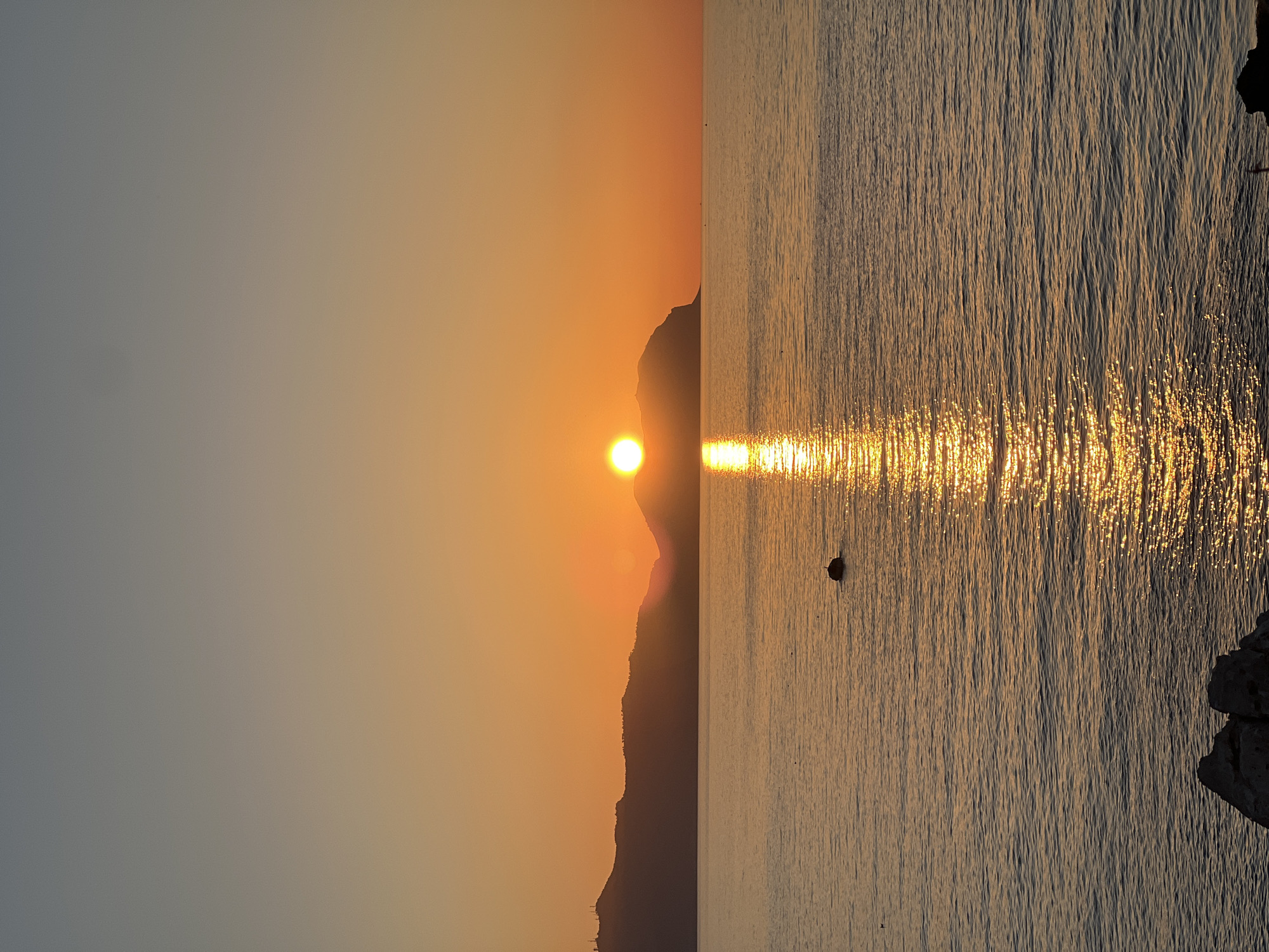 This image captures a sunset over the sea, with the sun positioned behind a silhouetted mountain range. The sky transitions from a pale orange near the horizon to a muted grey at the top. A bright reflection of the sun shimmers on the water's surface, creating a path of golden light. In the middle of the water, a dark, rounded object, possibly a buoy or a rock, is visible. In the foreground, dark, jagged rocks are partially submerged at the edge of the water. The overall scene suggests a peaceful coastal setting. While this specific image doesn't contain explicit indicators of Bagheria, Italy, it depicts a common coastal landscape found in the region, known for its picturesque seaside views and proximity to mountainous terrain.