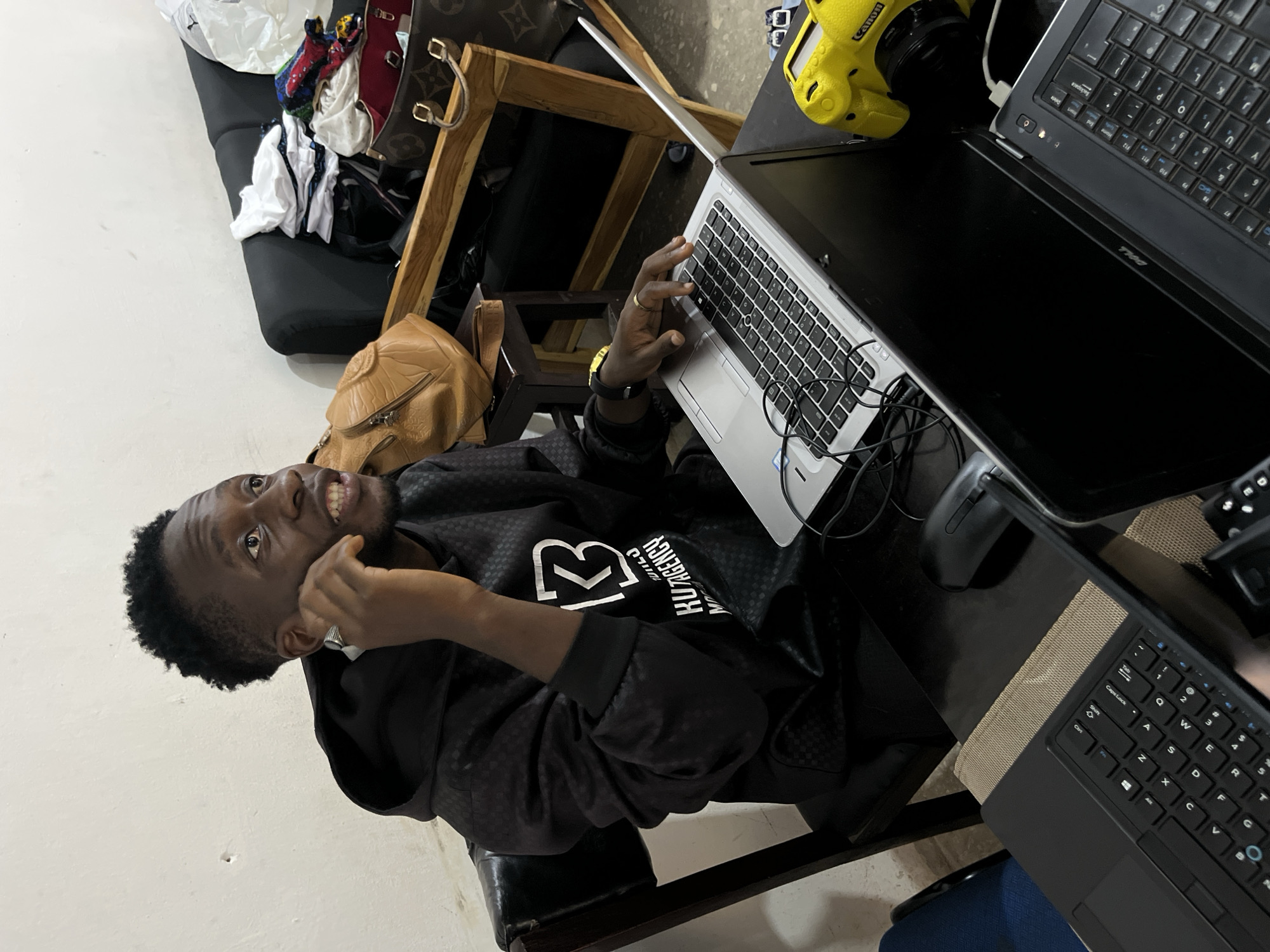 The image shows a young Black man sitting at a dark wooden table in what appears to be a room in Gura Top, Nigeria. He is wearing a black hoodie with a checkered pattern and a white logo on the chest. He is holding a phone to his right ear and looking upwards, with his mouth slightly open. In front of him are two laptops, one silver and one black, and a black computer mouse. To the right of the laptops is a bright yellow Canon camera. Behind the man, to the left, is a black leather chair. In the background, a black sofa is visible, piled with various items including clothing, handbags, and a white plastic bag. A wooden frame structure is positioned behind the sofa. The walls of the room are a neutral, light color, with some faint markings. The lighting appears to be artificial, casting shadows on the objects and the man. The overall impression is of a personal workspace or a room in a residence, possibly used for work or study.