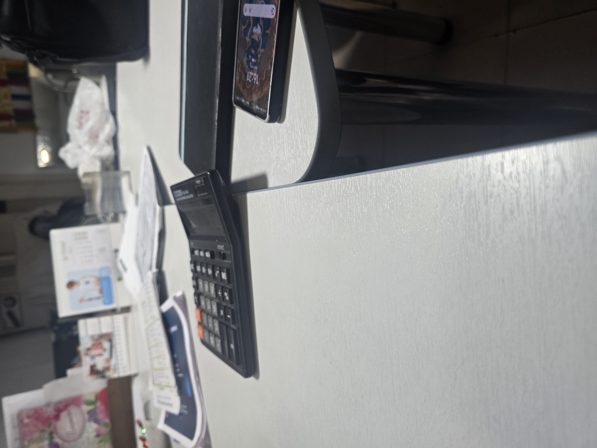 A close-up, eye-level shot captures a desk surface in Ulan-Ude, Russia. The surface, made of a white, textured material, is partially in shadow. On the desk, a black calculator with orange buttons is prominently featured. To the right, a smartphone with a dark case displays a colorful wallpaper with the time "16:21". The phone is positioned on a black rectangular object, possibly a stand or a tablet. In the background, slightly out of focus, are various desk items including papers, a calendar, and a black bag. The lighting suggests an indoor environment, possibly an office or home desk.
