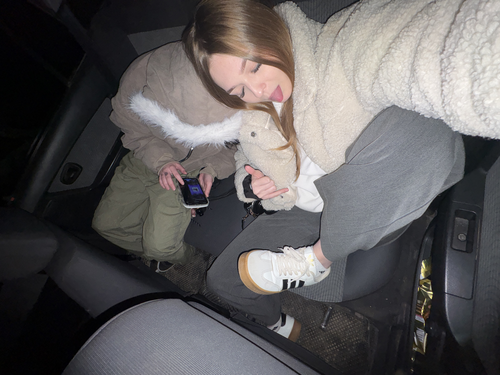 The image captures two young women relaxing in the backseat of a car at night in Chaikovsky, Russia. The scene is viewed from an elevated perspective, looking down into the vehicle's dark interior.
On the left, one woman wears a light brown or khaki puffer jacket with a white furry trim and green cargo pants. She is absorbed in her smartphone, holding it with both hands, with a small tattoo visible on her left hand. The phone's screen displays an active interface, possibly a music player or social media.
The second woman, on the right, leans against the first. She wears a cream-colored, fluffy teddy fleece jacket over a white top and dark grey sweatpants. Her head is tilted with her eyes closed, and she playfully sticks out her tongue. She holds a small, light-colored plush toy, possibly a sheep, in her left hand. Her footwear consists of white Adidas sneakers with distinctive black stripes and a gum sole.
The car's interior is dimly lit, with dark grey fabric seats and plastic panels. A gold-colored snack wrapper is visible on the floor next to the car door on the right side. The overall atmosphere is casual and relaxed, indicative of a casual social outing or journey, likely during the late hours given the pervasive darkness.