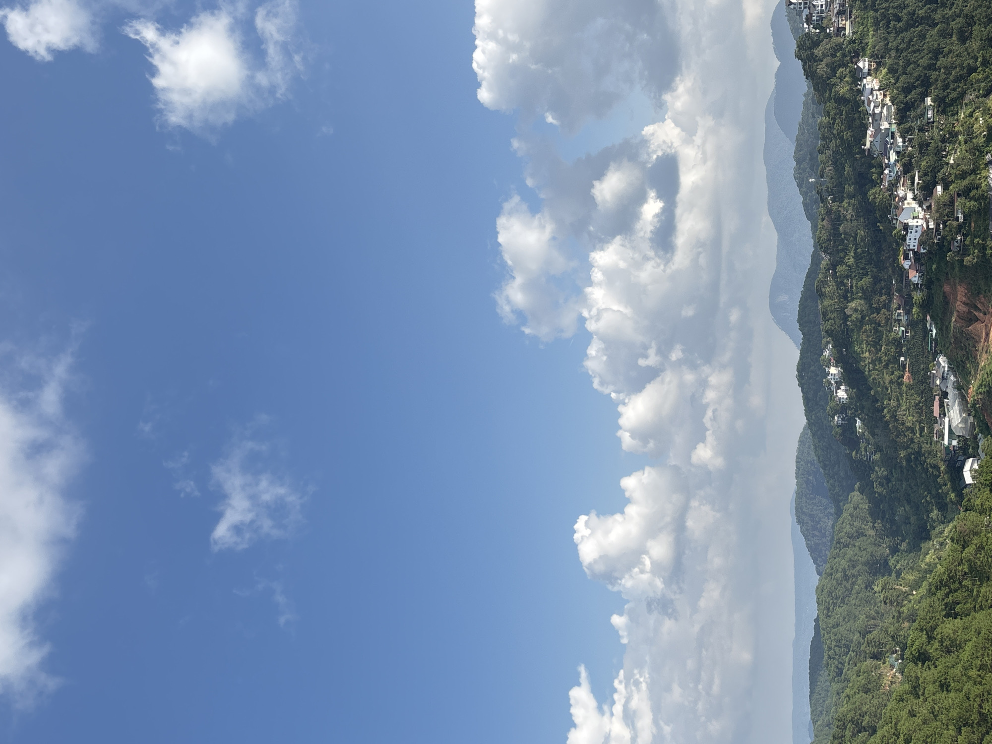 The image depicts a panoramic view of a lush, hilly landscape with scattered white buildings, characteristic of the city of Da Lat, Vietnam. The foreground is dominated by verdant green hills covered in dense foliage, likely pine trees, which are abundant in the region. The buildings are mostly white with red or grey roofs and appear to be situated on the slopes of these hills, forming small villages or residential areas that are nestled within the natural terrain.
In the midground, a series of undulating hills recede into the distance, their shapes softened by the atmospheric perspective. The furthest hills are a hazy blue, indicating their distance and the presence of mist or haze. The sky above is a vibrant blue, dotted with fluffy white cumulus clouds. Some of these clouds are large and dramatic, while others are smaller and wispy. The sunlight suggests it is daytime, likely late morning or early afternoon, given the brightness and the clarity of the sky.
The scene conveys a sense of tranquility and natural beauty, with the built environment harmoniously integrated into the mountainous landscape. The specific location within Da Lat is not explicitly identifiable from the image alone, but the architectural style of the buildings and the type of vegetation are strongly indicative of this Vietnamese highland city, renowned for its cool climate, pine forests, and French colonial architecture. There are no people or active events visible in the image. The focus is solely on the static landscape and the built structures within it.