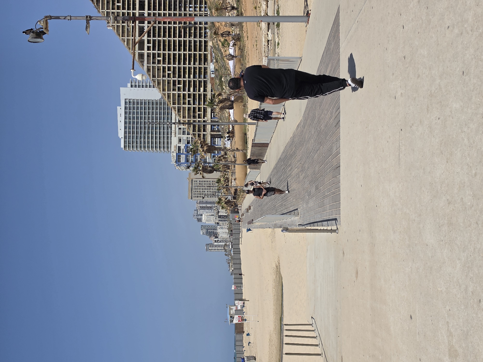 This image captures a bright, sunny day along the coastline of Bat Yam, Israel, showcasing a bustling promenade and beach alongside an urban skyline. The scene is dominated by a clear, vibrant blue sky.
In the foreground and midground, a wide promenade stretches horizontally, featuring both light-colored concrete and wooden plank sections. A man, dressed in a black t-shirt and black track pants with white stripes, stands on the concrete part of the promenade, facing away from the camera. His distinct shadow is cast on the ground, indicating strong sunlight. Further down the promenade, several other people are visible, walking or standing, some on the wooden section.
To the left of the promenade, a sandy beach unfolds, dotted with a few blue beach umbrellas and small structures, possibly lifeguard stands or beach facilities. Beyond the beach, the calm, blue waters of the Mediterranean Sea extend into the distance.
The right side of the image reveals the city skyline of Bat Yam. A prominent high-rise building with an exposed concrete skeleton suggests ongoing construction. Several other modern, white multi-story buildings, interspersed with palm trees, complete the urban backdrop. A tall metal lamp post with a large light fixture stands prominently on the promenade's edge on the right. A set of concrete stairs with a metal railing provides access from the promenade down to the beach.
The overall scene suggests a pleasant, active day in a coastal city environment. There is no visible text in the image.