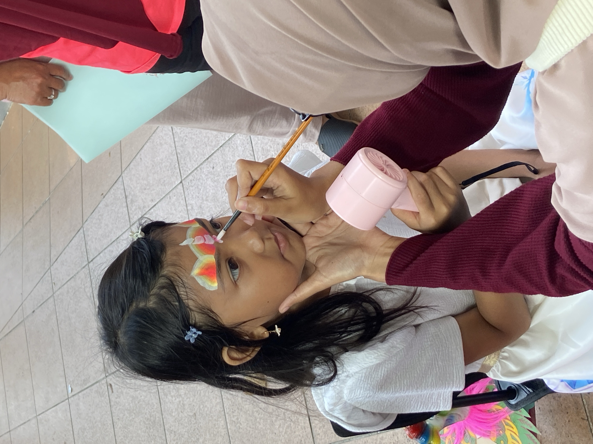 A young girl is sitting down while a person applies face paint with a small brush. The face paint consists of yellow, orange, red, and white colors, and appears to be a butterfly design near her eye. Another person is holding a small pink fan near the girl's face, possibly to dry the paint. The girl has dark hair with small blue clips. The person painting her face is wearing a maroon shirt. The scene is likely in a public space, as indicated by the tiled floor. The location is Surabaya, Indonesia.
