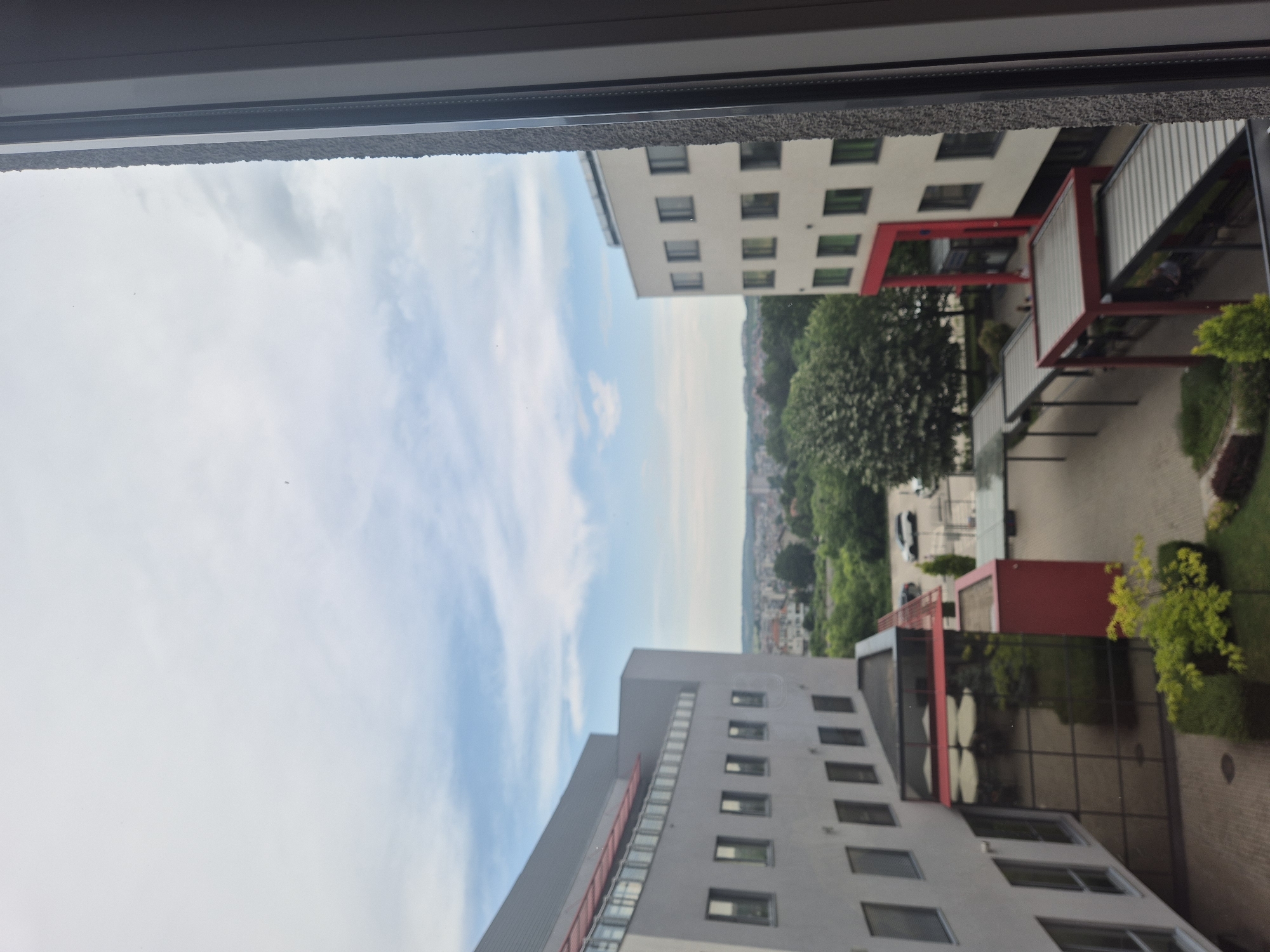 An elevated view captured from a window displays a modern urban landscape under a partly cloudy sky. The right edge of the image is occupied by a light-colored window frame, showing its textured exterior and smooth interior components. Two prominent multi-story buildings, characterized by light grey facades and numerous dark rectangular windows, frame a central courtyard.
The building on the left features a red, railed balcony structure near its upper edge. Lower down, a large glass-enclosed section with a red support structure houses what appears to be a cafe or restaurant, with multiple light-colored outdoor umbrellas and indistinct figures of people visible inside. The building on the right also exhibits red architectural detailing, including vertical and horizontal beams on its lower facade.
The courtyard between the buildings is paved with light-colored interlocking bricks. Several covered walkways, featuring red frames and light-colored ribbed roofs, extend from the right building across the courtyard. Indistinct human figures are present on these walkways. The courtyard also contains manicured green landscaping, including hedges and small trees with bright green foliage. In the background of the courtyard, a paved area holds at least two parked vehicles, including a white SUV. Beyond the immediate structures, a sprawling cityscape with numerous buildings extends towards a distant horizon line, where a range of green hills or mountains is visible under the blue sky with scattered white clouds. The scene suggests a daytime setting with good visibility.