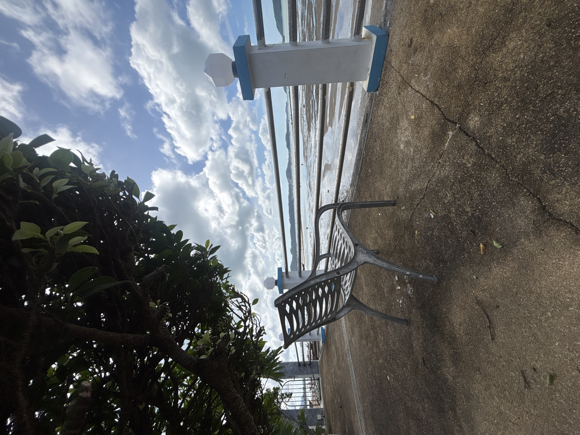 An empty metal bench sits on a concrete walkway overlooking a body of water in Rawai, Thailand. The bench faces away from the camera, towards the water. A railing with a white post topped with a white light fixture runs along the edge of the walkway. A large tree with dark branches and green leaves partially shades the foreground. The sky above is blue with white, puffy clouds. The water appears to be at low tide, exposing a muddy, wet shoreline. In the distance, hills and possibly other islands are visible. The concrete walkway is cracked in several places. There are no people or animals visible in the image.