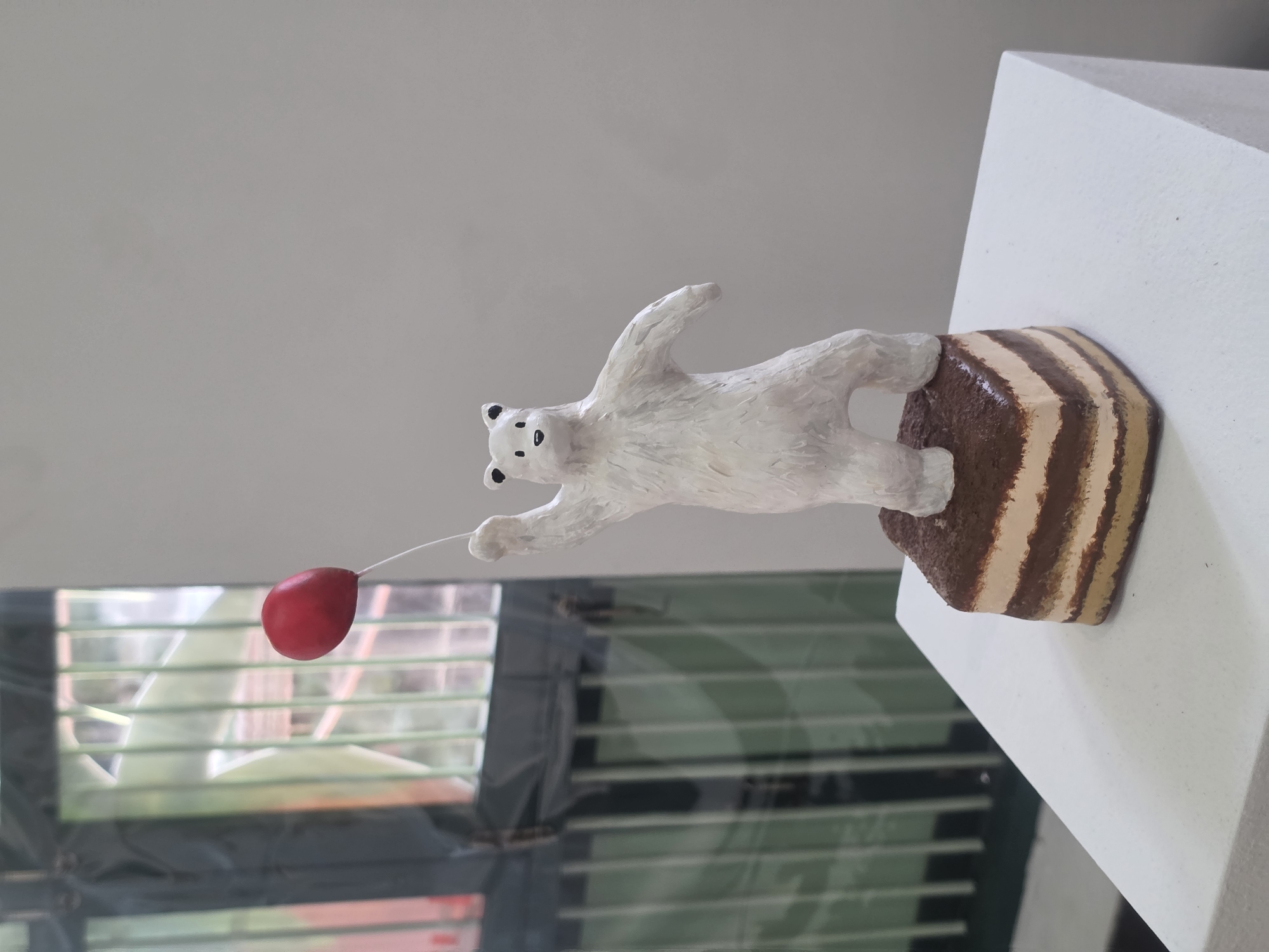 A hand-crafted polar bear figurine stands on a small, striped cake-shaped pedestal. The figurine is white, with black markings for the eyes and nose, and appears to be reaching towards a small, red balloon held by a thin string. The figurine and pedestal rest on a white, possibly plaster, surface, likely indoors. The background includes a window with venetian blinds, the exterior is nondescript, it is not possible to establish the location in Bangkok, Thailand.