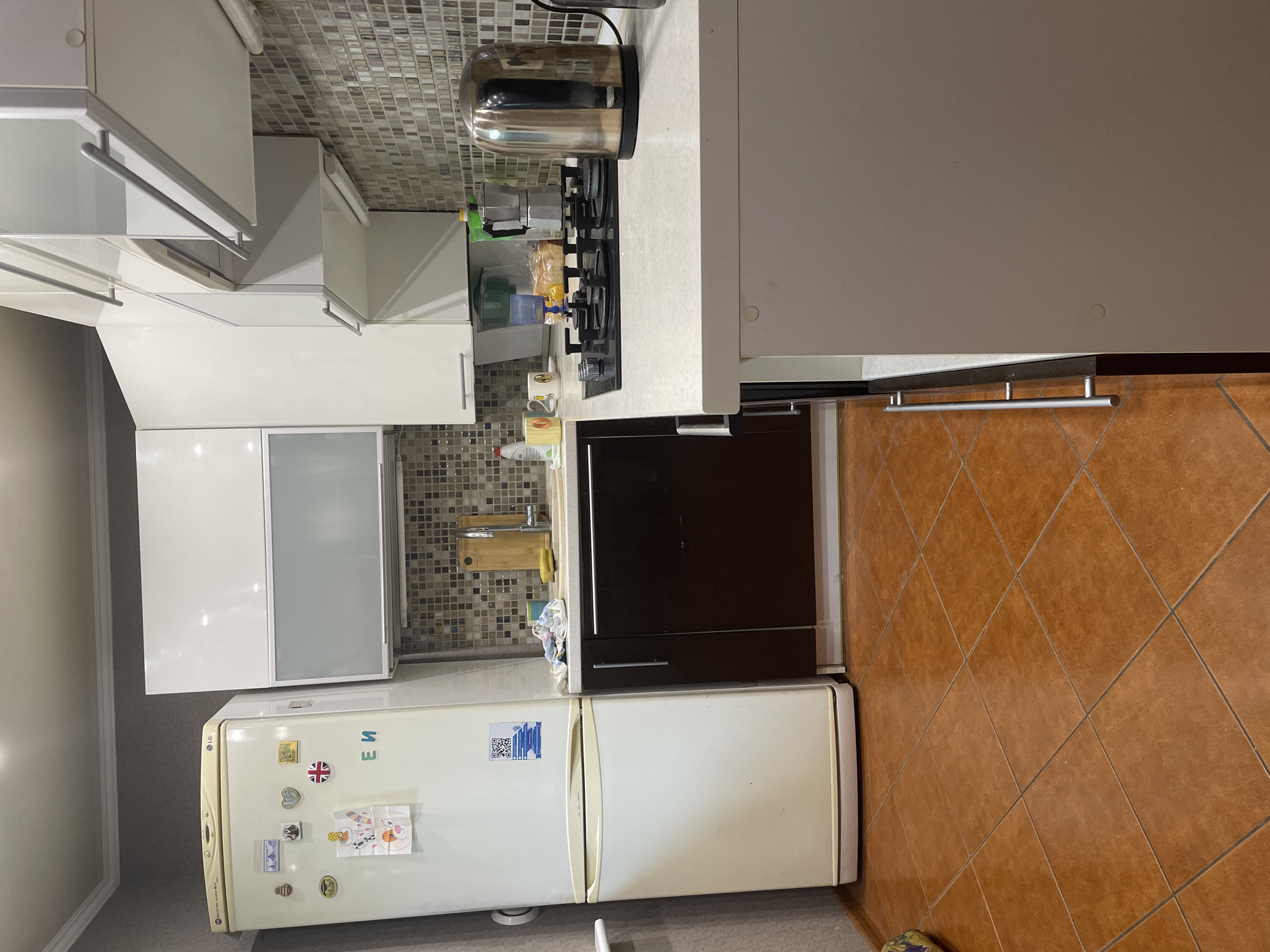 A medium shot captures a domestic kitchen environment. A white LG refrigerator stands on the left, adorned with various magnets and stickers, including what appears to be a QR code. To its right, a dark brown cabinet is visible, adjacent to a speckled tiled backsplash that runs along the wall. Above the cabinets, white upper cabinets with frosted glass inserts are mounted. A countertop with a stovetop and a stovetop kettle is positioned in the foreground on the right. The floor is tiled with reddish-brown square tiles. The scene is illuminated by artificial lighting. The image was taken in Kaliningrad, Russia.