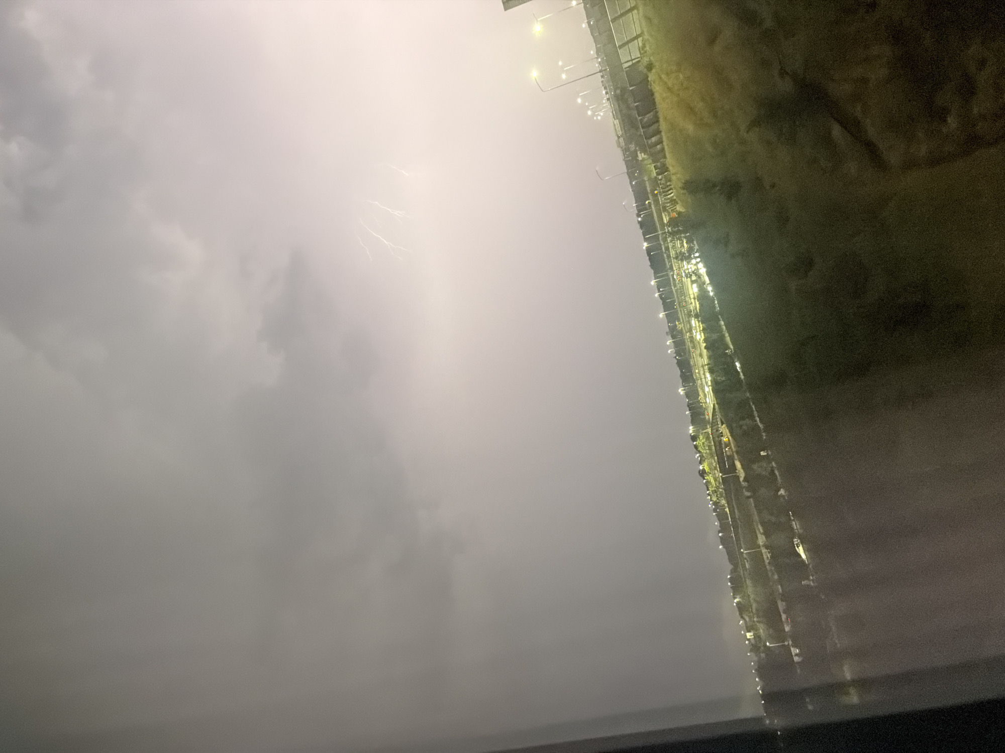 A nighttime thunderstorm illuminates the sky over the A9 highway and surrounding areas in Badhoevedorp, Netherlands. Jagged streaks of lightning flash against a backdrop of dark, ominous clouds. Streetlights line the highway, casting a warm glow on the road and the adjacent grassy slopes. In the foreground, a dimly lit window frame creates a slight obstruction, suggesting the photo was taken from indoors, possibly a residential building or hotel. The overall scene captures a dramatic natural event occurring within an urban landscape.
