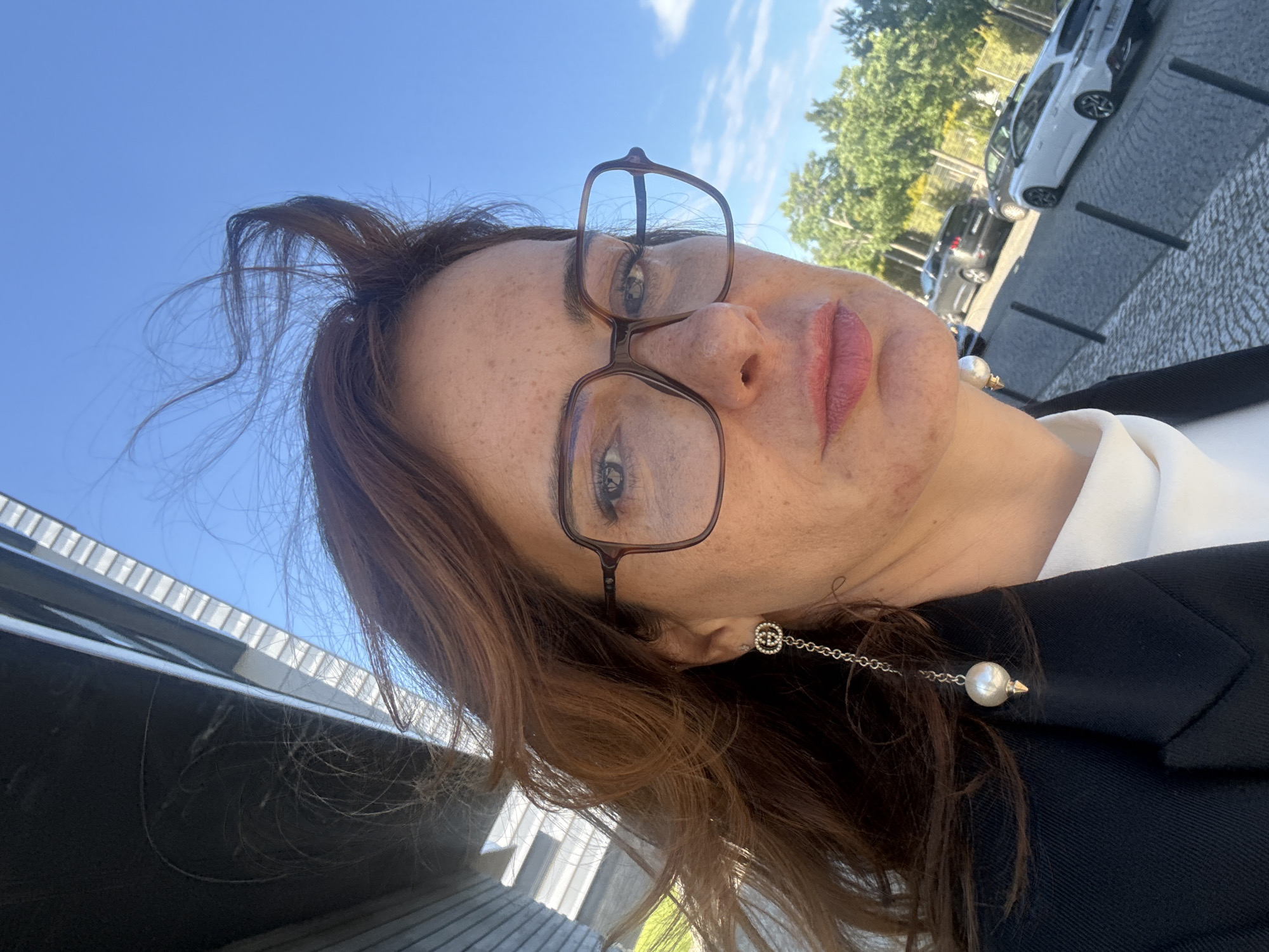 A woman is taking a selfie, looking up towards the camera. She has light skin, brown hair, and is wearing eyeglasses with brown frames. She is also wearing an earring that consists of a small stud attached to a chain with a pearl at the end. She is wearing a black jacket over a white shirt. The background includes a blue sky, trees, parked cars, and the corner of a building. The location appears to be in an urban area, possibly in Loures, Portugal.