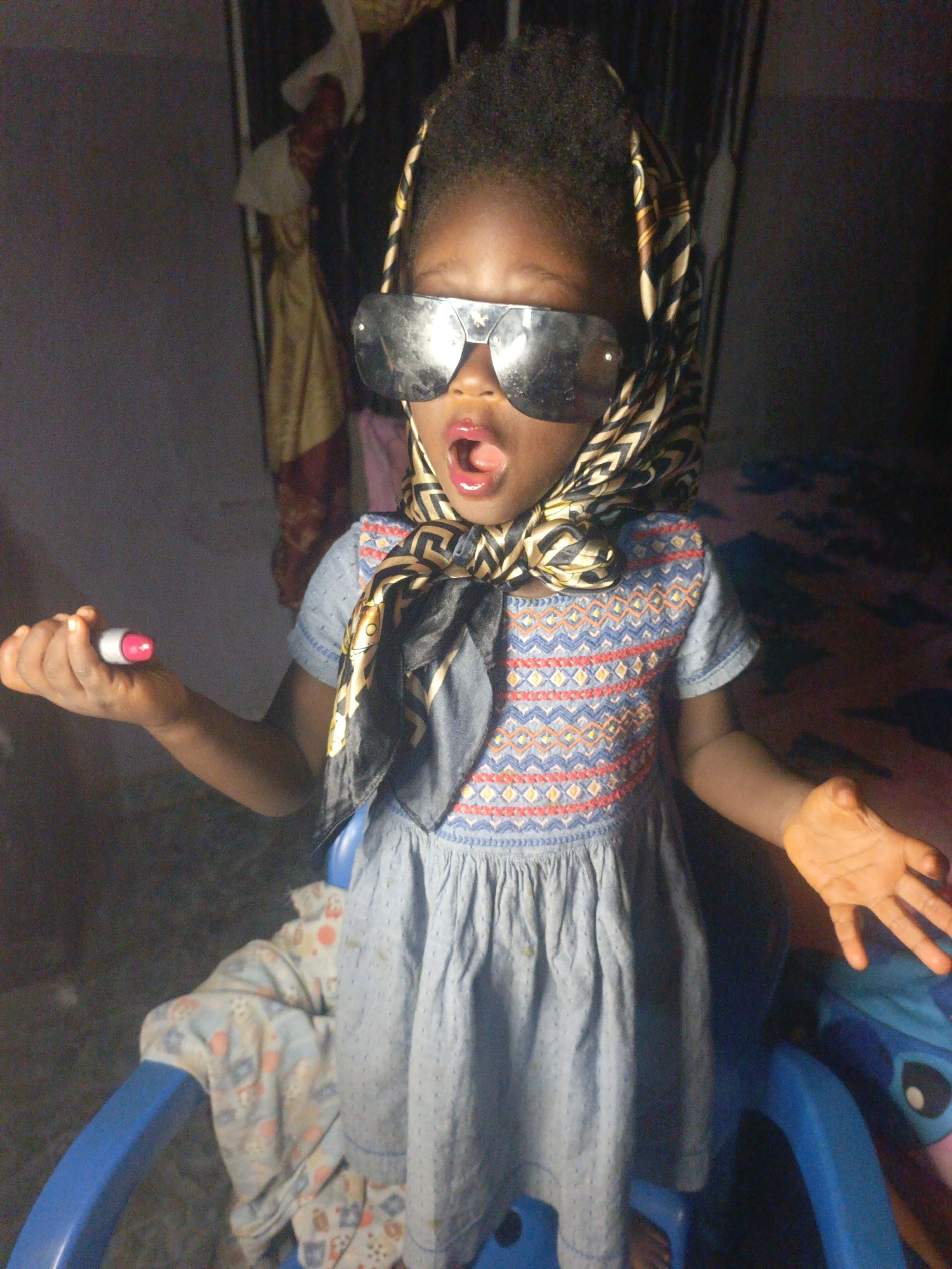 A child, likely a toddler, is seated in a blue plastic chair in Akwanga, Nigeria. The child is wearing sunglasses, a patterned headscarf, and a blue dress with embroidered details on the bodice. The child's mouth is open. The child is holding a lipstick in one hand and has orange residue on the other hand. A patterned blanket is draped over the chair. In the background, there is a wall with a poster or artwork hanging on it. The lighting is dim.