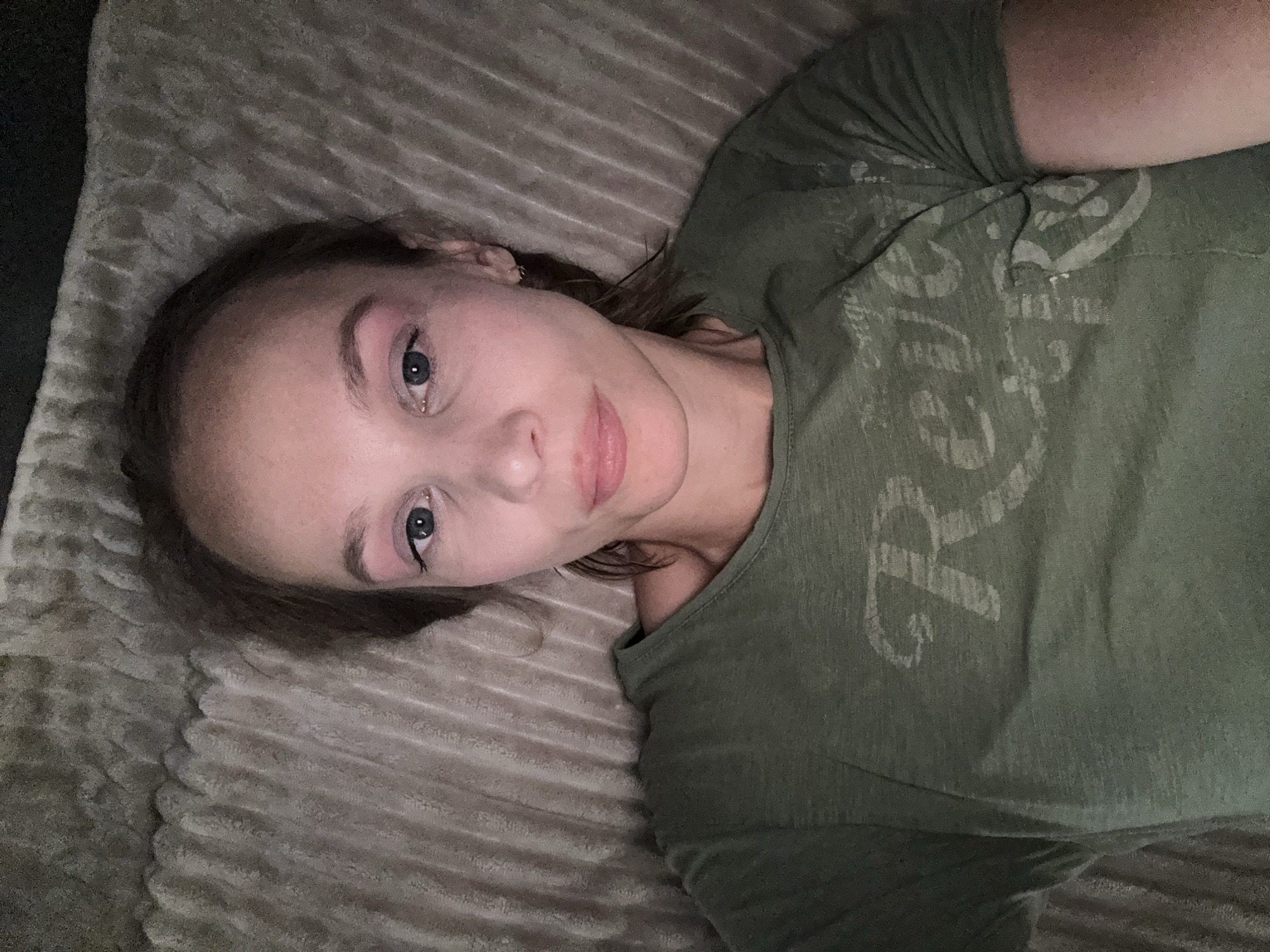 A young Caucasian woman is lying down on a textured surface that appears to be a bed or sofa. She is wearing a green t-shirt with a distressed graphic that reads "Reve R". The woman has light skin, blue eyes, and light brown hair. Her eyelids are adorned with pink eyeshadow and black eyeliner. The background is a neutral-toned, textured fabric. The scene is dimly lit, suggesting it may be nighttime or an indoor setting with low light. The photograph is taken from a high angle, looking down at the woman. The specific location in the city of Überherrn, Germany, cannot be determined from the visual information provided in the image.
