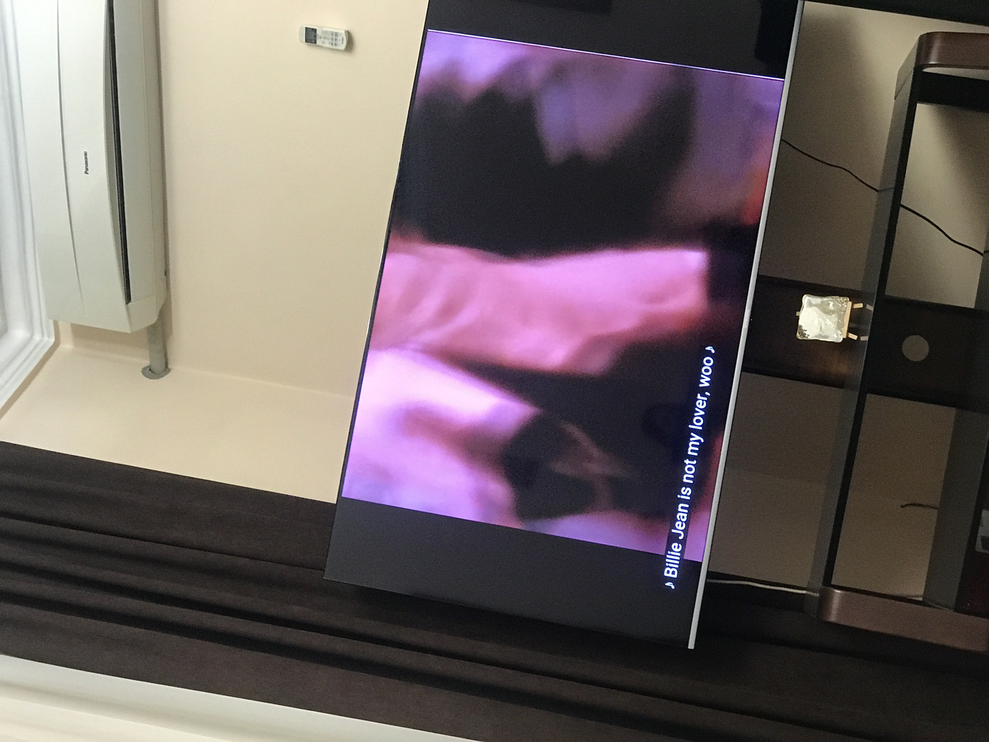 A television screen displaying a video with the caption "Billie Jean is not my lover, woo" is situated in an interior space. An air conditioning unit, bearing the brand "Panasonic," is mounted on the wall. A remote control is affixed to the same wall. A small, transparent object is visible on a nearby shelf. The presence of a dark-colored surface suggests the lower part of the television is positioned on a piece of furniture. The location within Yerevan, Armenia is not evident from the visual information provided.