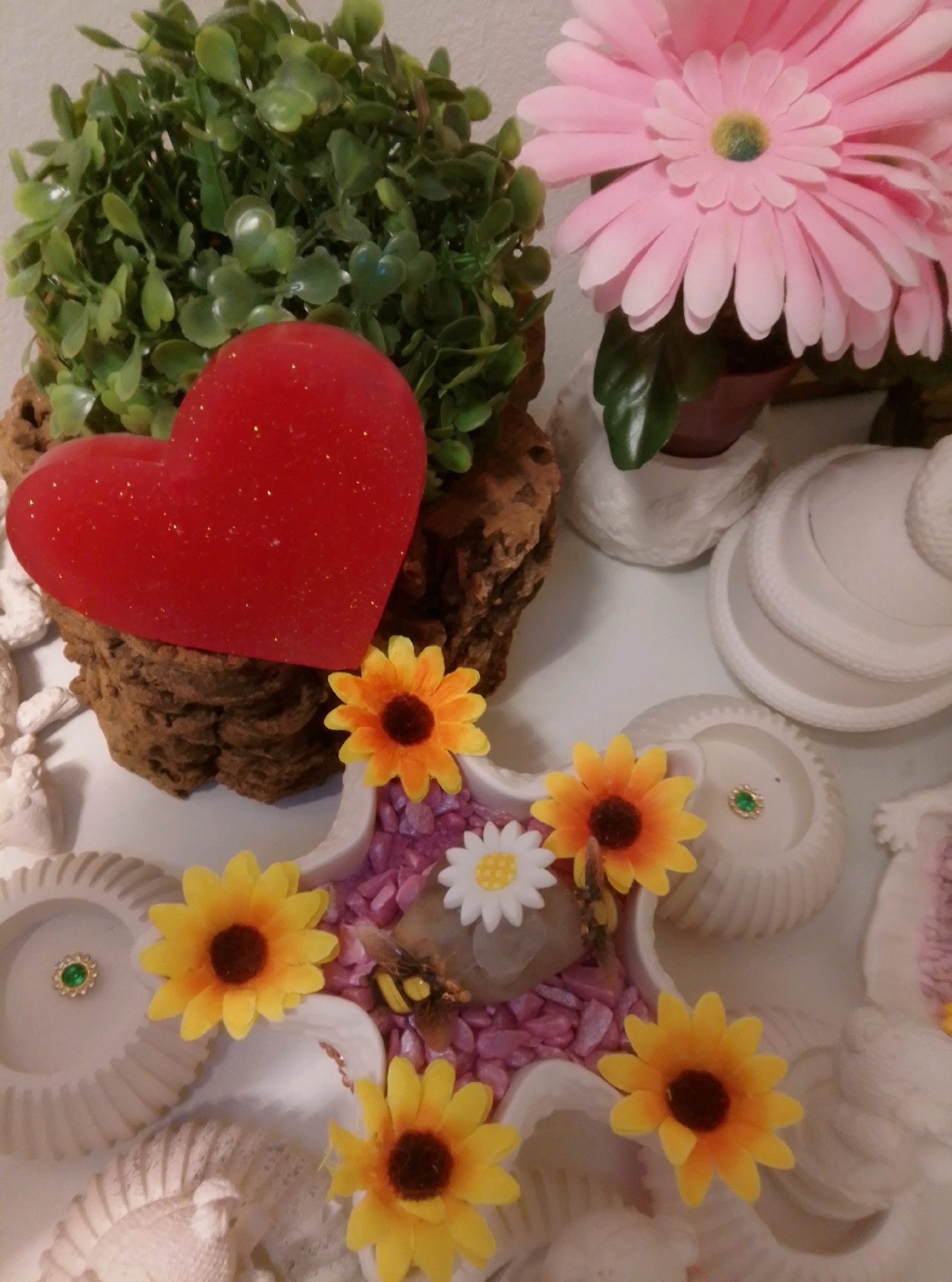 The image depicts a close-up view of a decorative display, likely an indoor arrangement. In the upper left, a bright red, glitter-covered heart-shaped object rests against a textured, brown, bark-like base. Behind the heart is a cluster of small, green, artificial leaves. To the right of the heart, a large, pale pink artificial daisy with a yellow-green center is prominently displayed.
Below the heart and bark base, the scene transitions to a collection of small, artificial sunflowers with yellow petals and dark brown centers. These flowers are arranged around small, white, decorative bowls or holders, some of which contain small, pink, decorative stones. One of these white holders has a small, green jewel or embellishment. A small, artificial bee is also visible amongst the flowers and stones. The overall presentation suggests a themed tabletop arrangement or craft project. There is no indication of the city of Wedel, Germany, in this image, nor are there any people, actions, or interactions depicted.