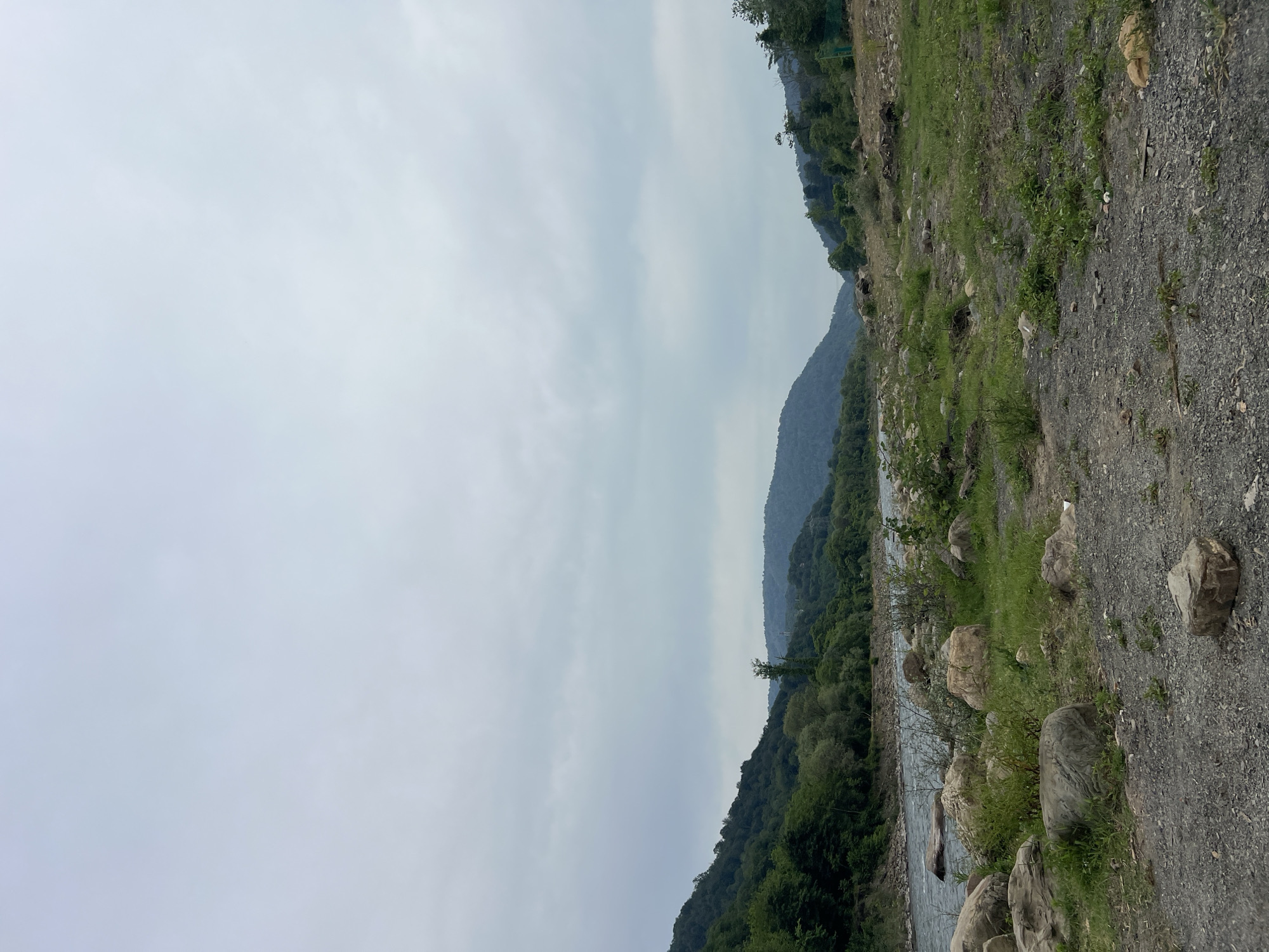 The image shows a scenic view of a river flowing through a valley with mountains in the background. The sky is overcast with clouds. The foreground is rocky and covered with sparse grass. The mountains are densely forested with green trees. The river is wide and appears to be flowing calmly. This scene is likely located in the outskirts of Sochi, Russia, known for its natural beauty and mountainous terrain.